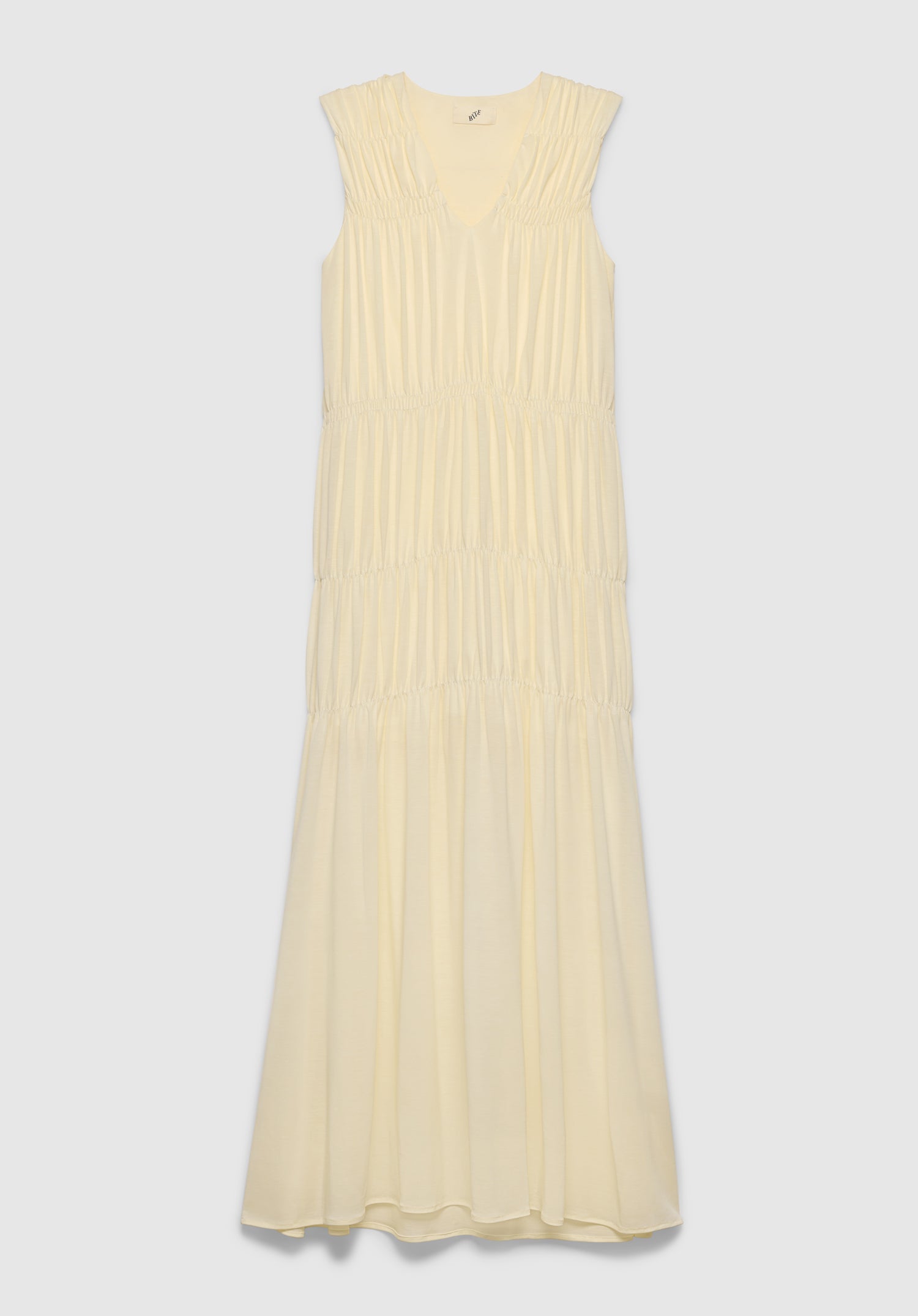 A light cream sleeveless maxi dress with a V-neckline and a tiered, gathered design, featuring soft pleating and a flowing silhouette, displayed against a plain background.
