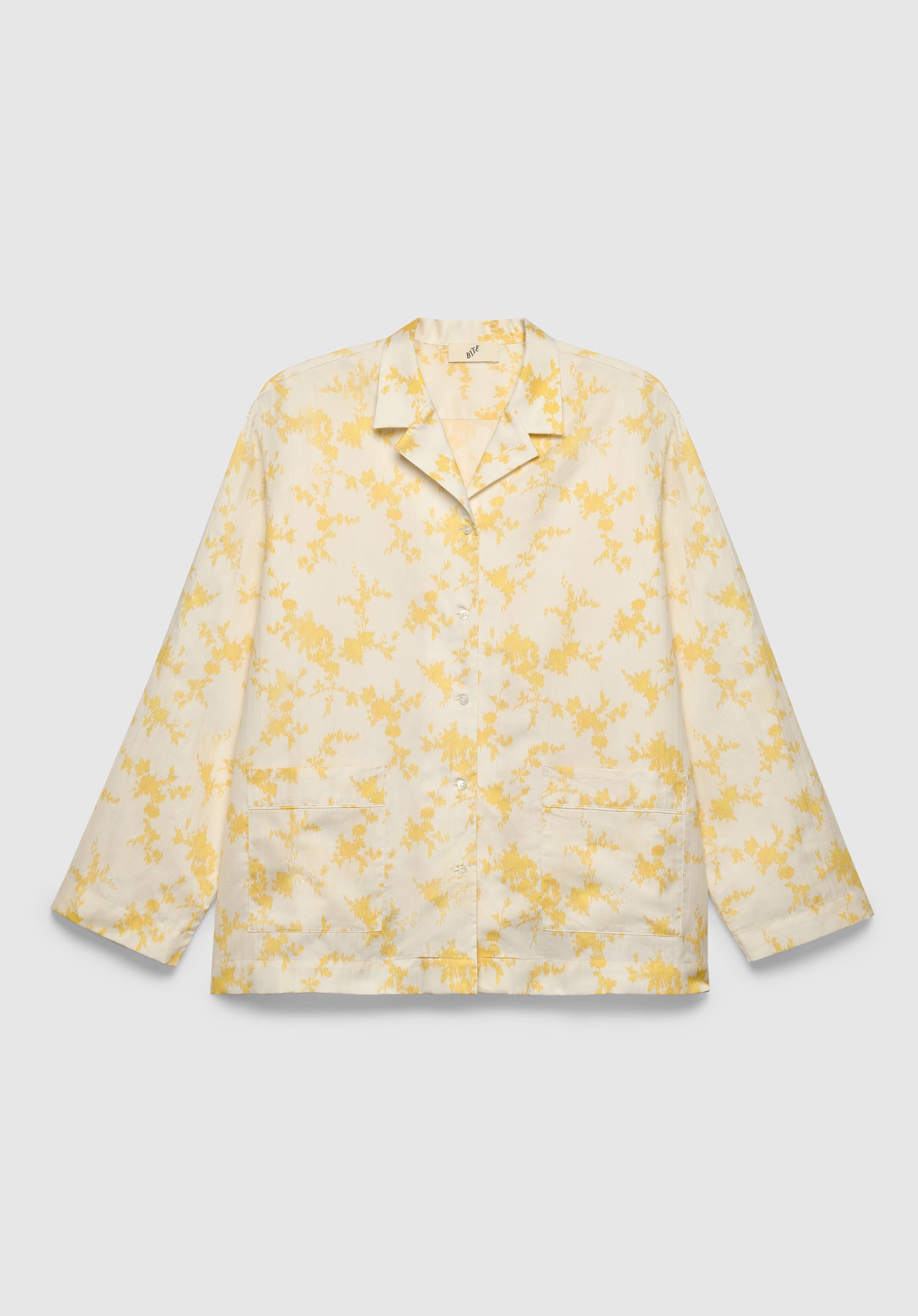 A cream-colored long-sleeve shirt with a relaxed fit, featuring a notched collar, front button closure, patch pockets, and a delicate yellow floral print, displayed against a plain background.

