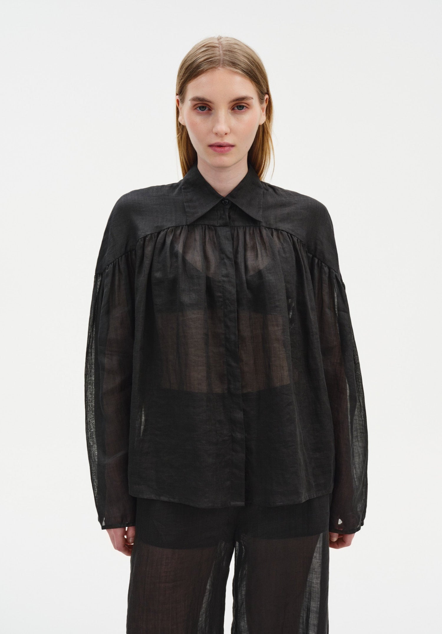 Front view of a model wearing a sheer black long-sleeve blouse with a pointed collar and gathered detailing, paired with matching translucent trousers, emphasizing the lightweight, semi-transparent fabric and relaxed silhouette.
