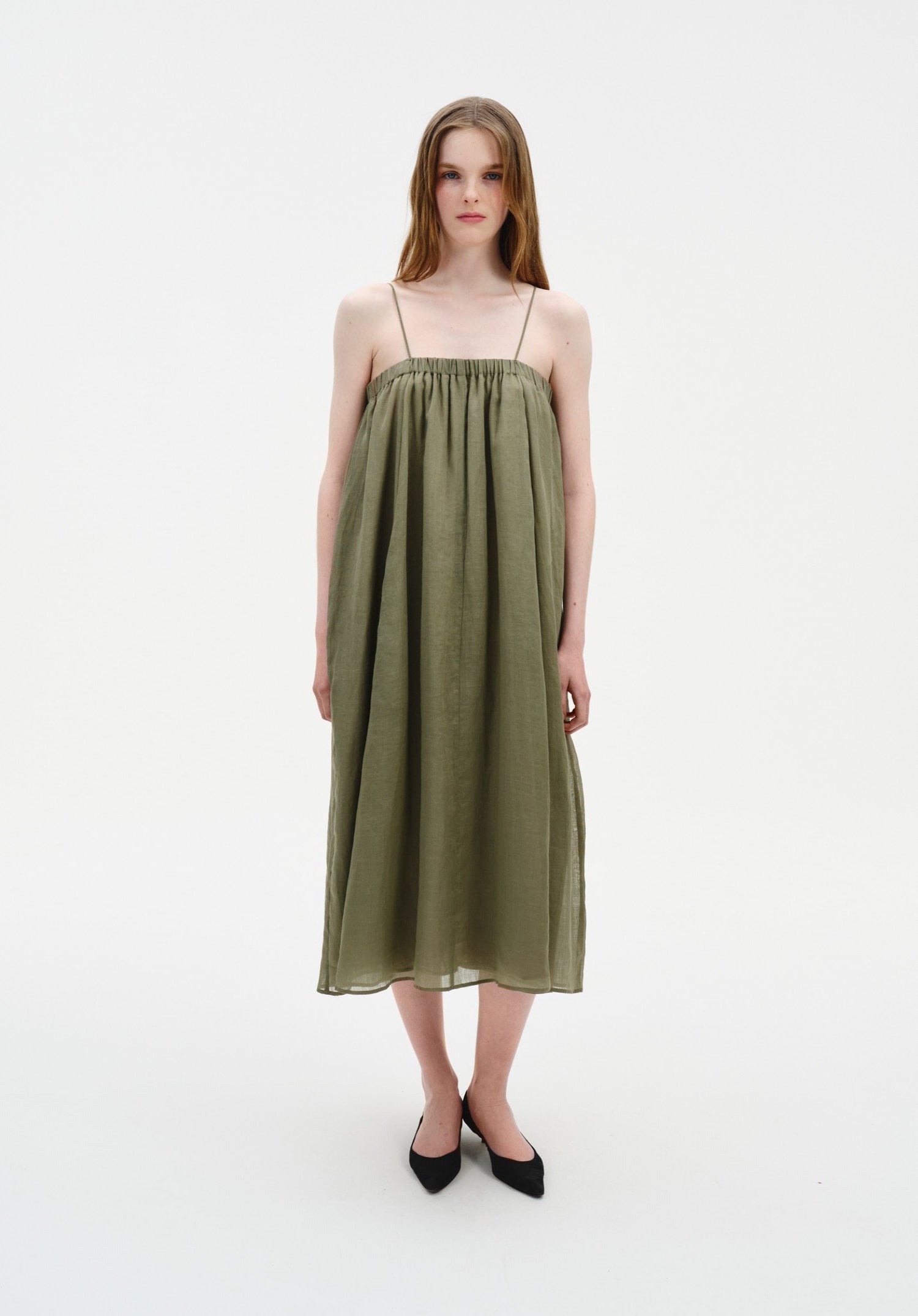 A woman stands facing forward in a straight, centered stance with feet close together, wearing an olive-green sleeveless midi dress with thin straps and a loose, gathered silhouette, paired with black pointed heels.
