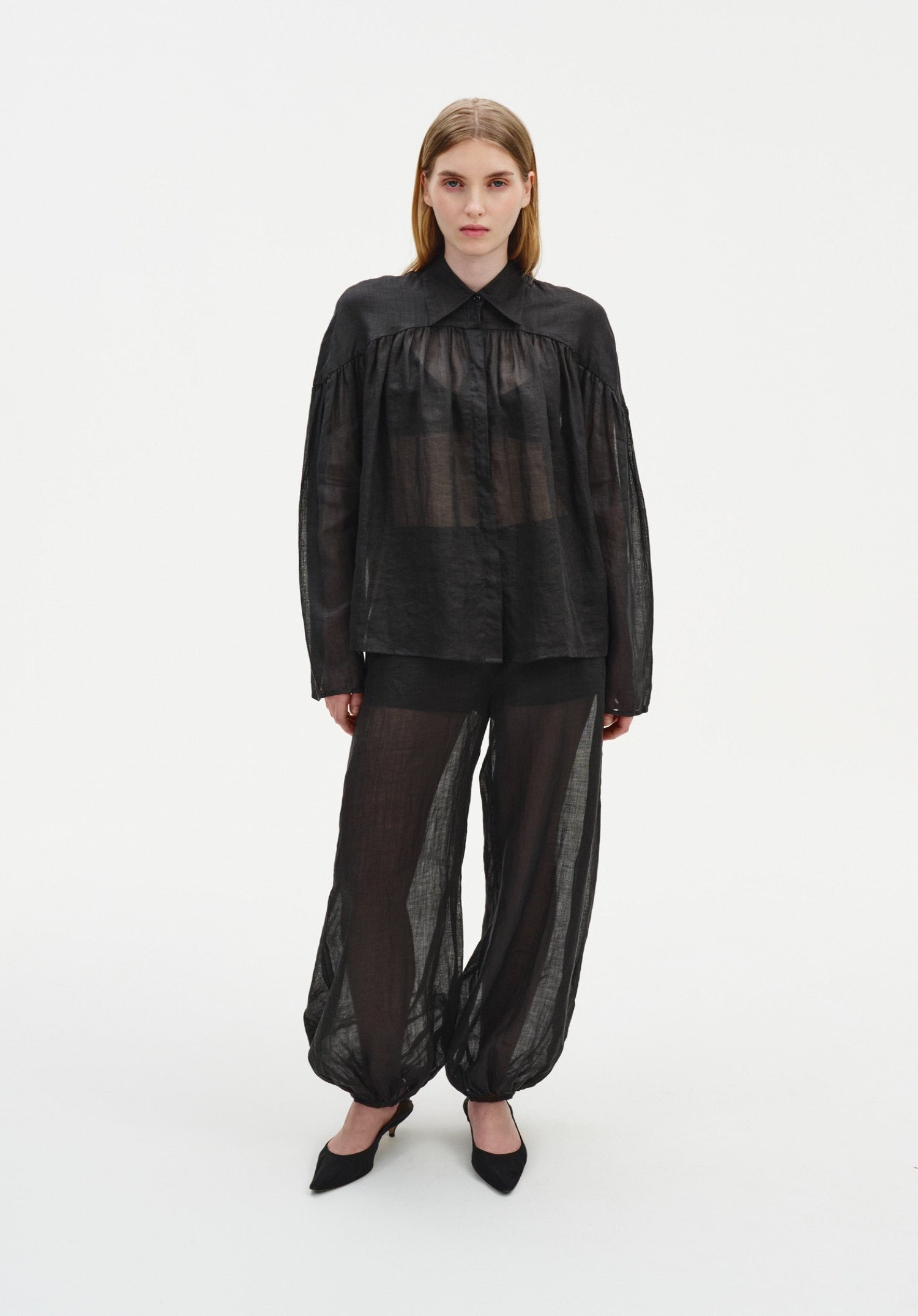 A woman stands facing forward against a plain white background, wearing a sheer black long-sleeve blouse with a collar and gathered detailing, paired with matching semi-transparent billowy trousers and black pointed heels.
