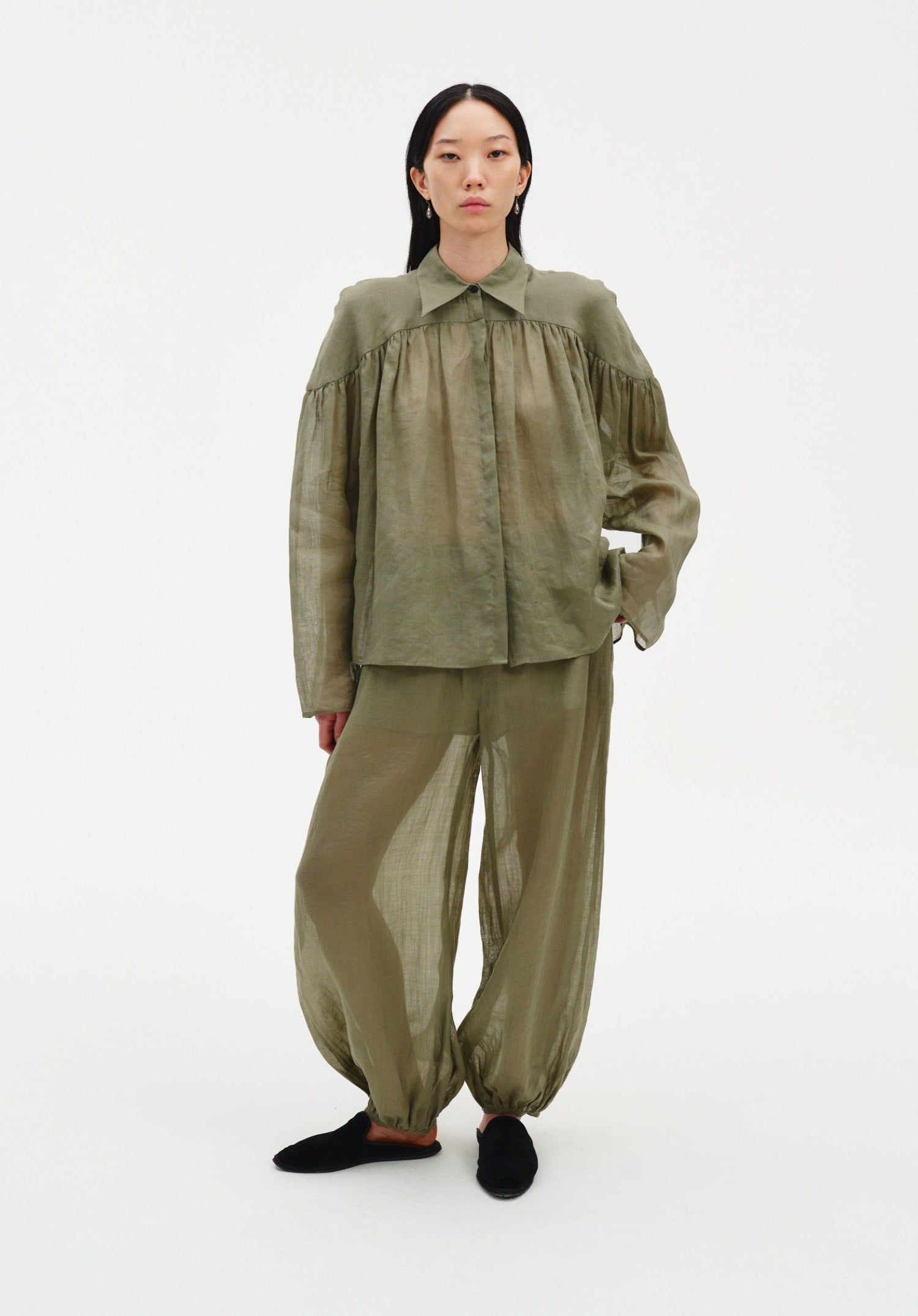 A woman stands facing forward in a relaxed stance against a plain white background, wearing an olive-green sheer blouse with a pointed collar and gathered detailing across the chest, paired with matching lightweight, billowy trousers and dark slip-on shoes.
