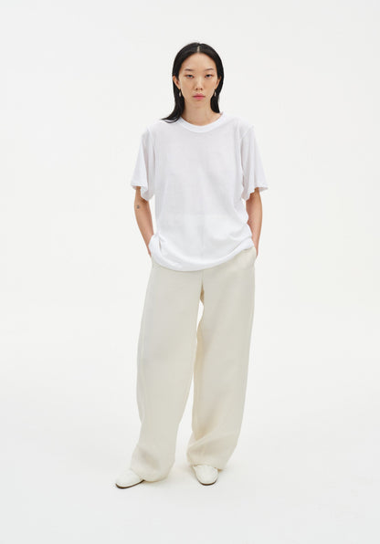 A woman stands facing forward in a relaxed stance against a plain white background, wearing an oversized white T-shirt and loose cream-colored wide-leg trousers with light-colored shoes, her hands in her pockets and posture slightly casual.
