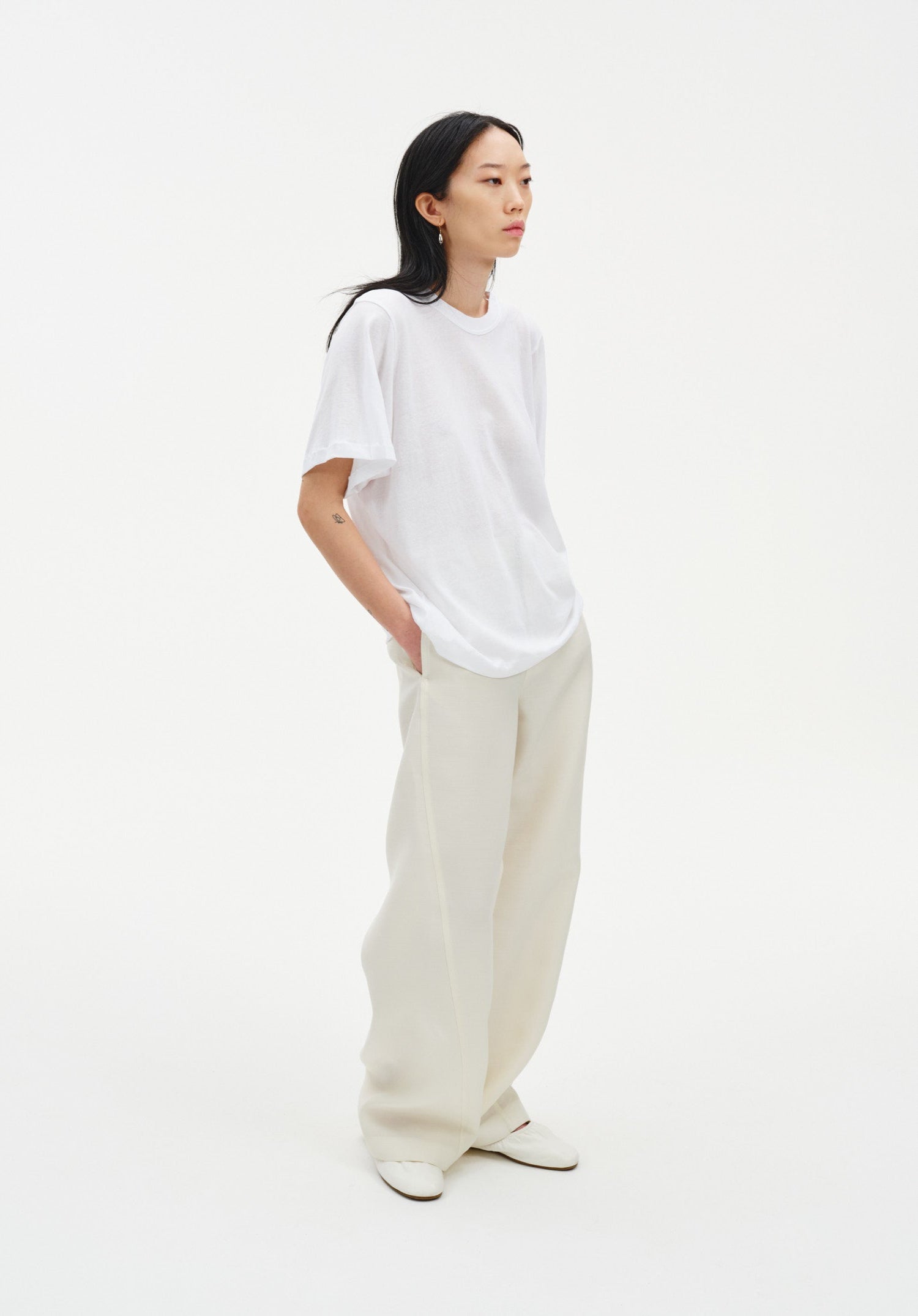 A woman stands in a relaxed three-quarter side pose against a plain white background, wearing an oversized white T-shirt tucked into loose cream-colored trousers and light-colored shoes. Her dark shoulder-length hair is worn down, and she looks slightly off to the side with her hands in her pockets.