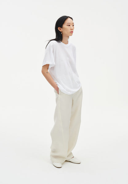 A woman stands in a relaxed three-quarter side pose against a plain white background, wearing an oversized white T-shirt tucked into loose cream-colored trousers and light-colored shoes. Her dark shoulder-length hair is worn down, and she looks slightly off to the side with her hands in her pockets.