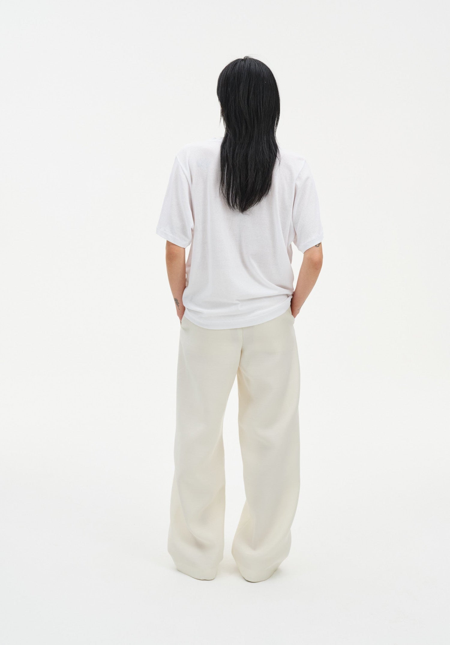 Rear view of a woman standing against a plain white background, wearing a relaxed white short-sleeve top and loose cream-colored trousers. Her dark shoulder-length hair falls straight down her back, and her hands rest in her pockets.
