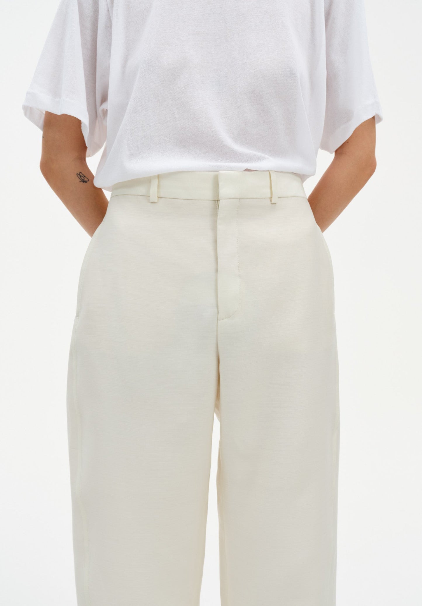 Close-up of cream-colored tailored trousers with a clean waistband and front seam detail, paired with a white top tucked in, highlighting the smooth fabric and structured fit.
