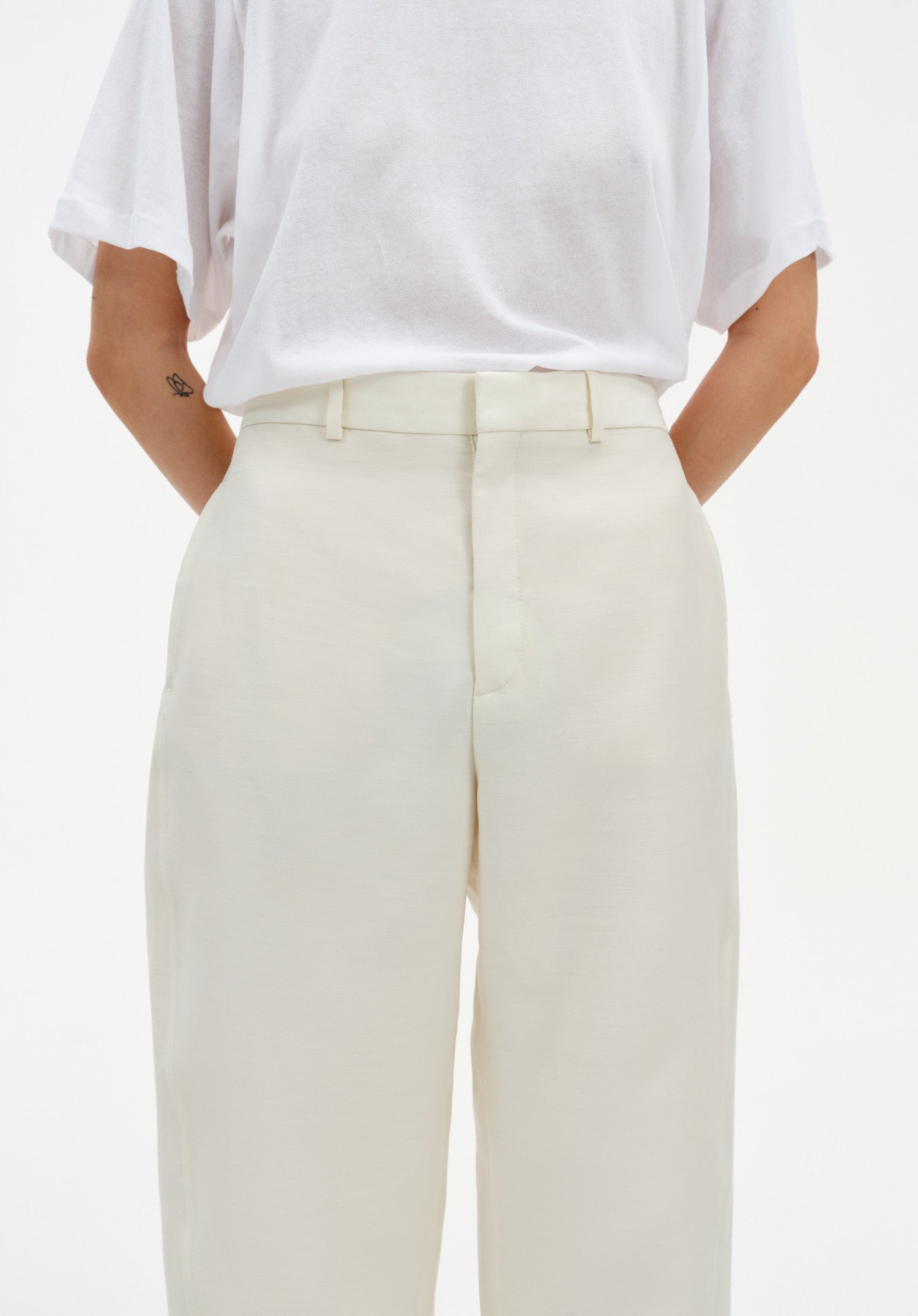 Close-up of cream-colored tailored trousers with a clean waistband and front seam detail, paired with a white top tucked in, highlighting the smooth fabric and structured fit.
