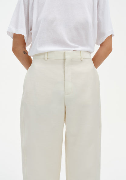 Close-up of cream-colored tailored trousers with a clean waistband and front seam detail, paired with a white top tucked in, highlighting the smooth fabric and structured fit.
