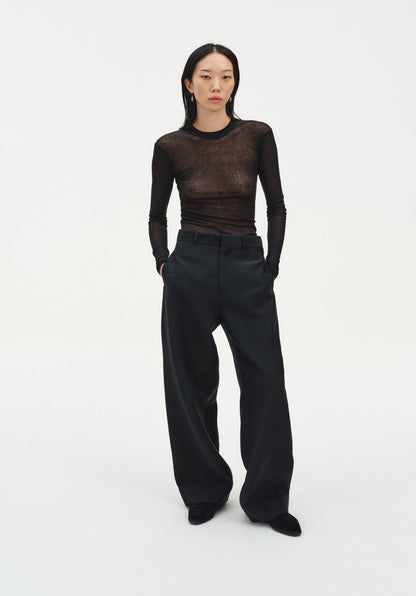 A woman stands facing forward in a relaxed stance with a slight weight shift, hands in her pockets, against a plain white background. She wears a sheer long-sleeve dark top and black wide-leg trousers with black shoes, her dark shoulder-length hair worn down and her expression neutral.
