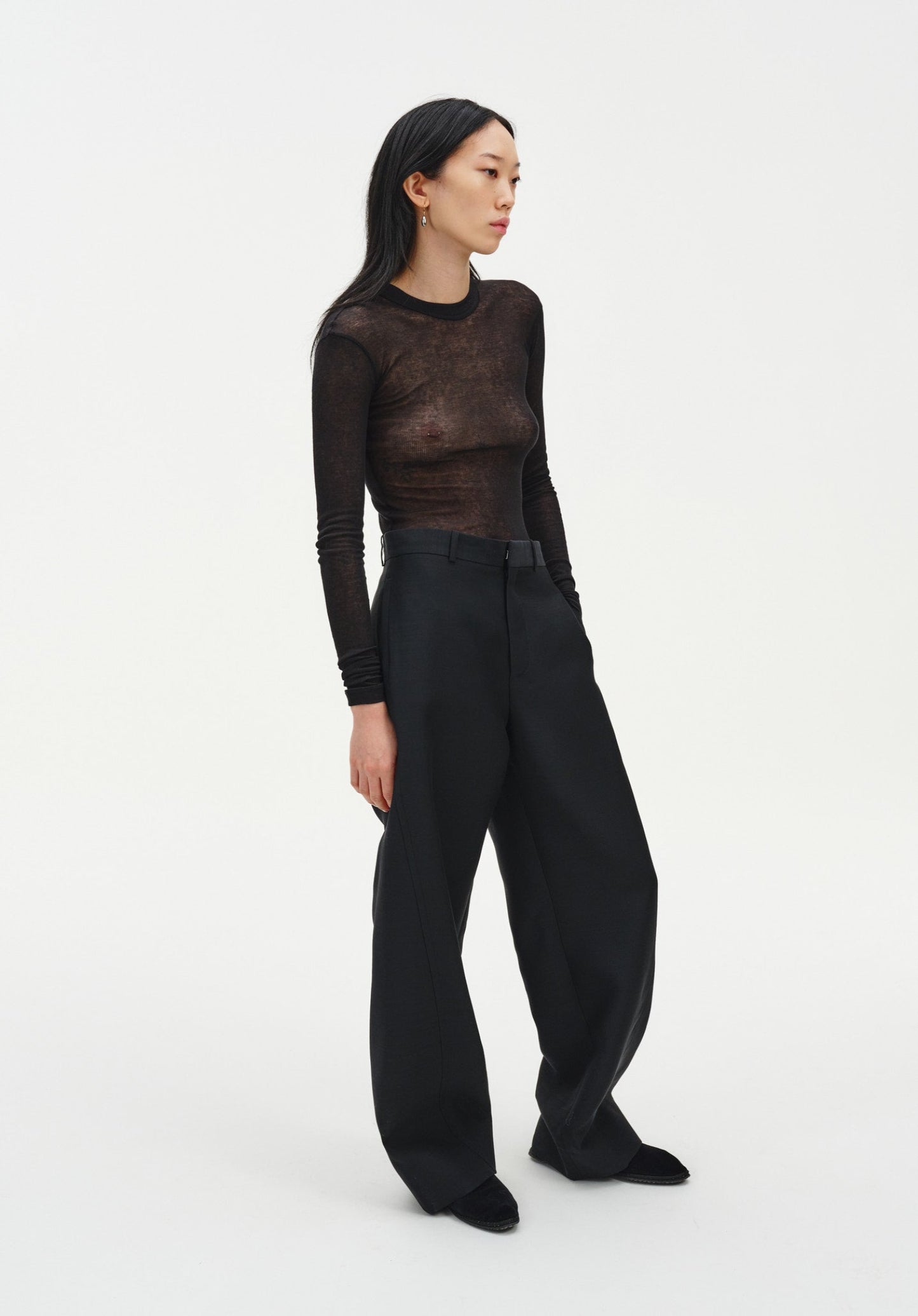 A woman stands in a relaxed three-quarter side pose against a plain white background, wearing a sheer long-sleeve dark top and black wide-leg trousers with black shoes. Her dark shoulder-length hair is worn down, and she looks slightly to the side with a neutral expression, one hand resting in her pocket.
