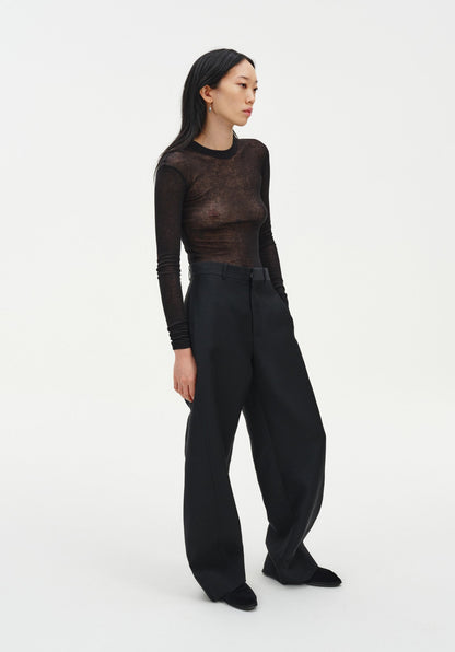 A woman stands in a relaxed three-quarter side pose against a plain white background, wearing a sheer long-sleeve dark top and black wide-leg trousers with black shoes. Her dark shoulder-length hair is worn down, and she looks slightly to the side with a neutral expression, one hand resting in her pocket.
