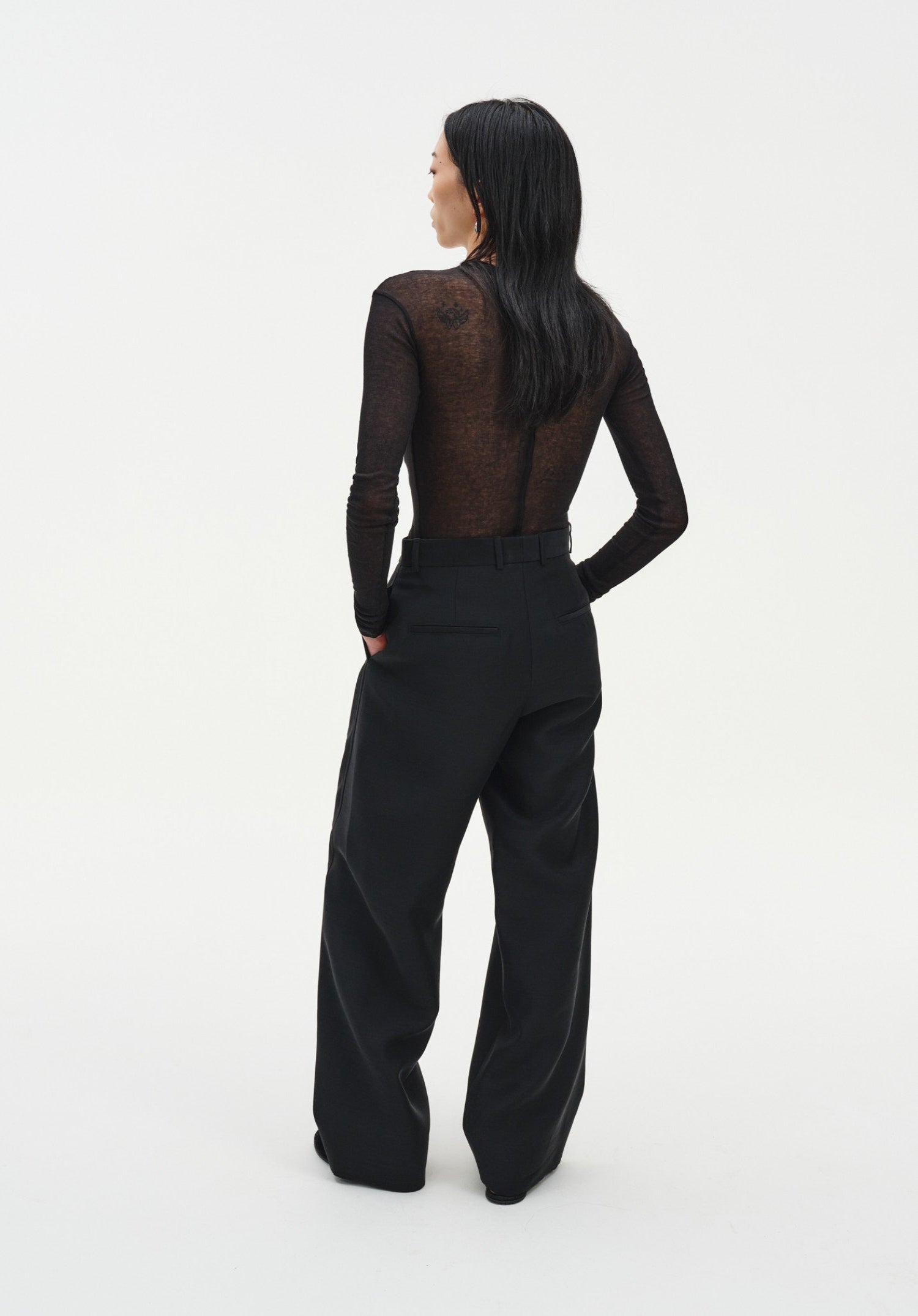 Rear three-quarter view of a woman against a plain white background, wearing a sheer long-sleeve dark top tucked into black tailored trousers. Her dark shoulder-length hair falls down her back, and one hand rests in her pocket, highlighting the relaxed fit and clean lines of the outfit.
