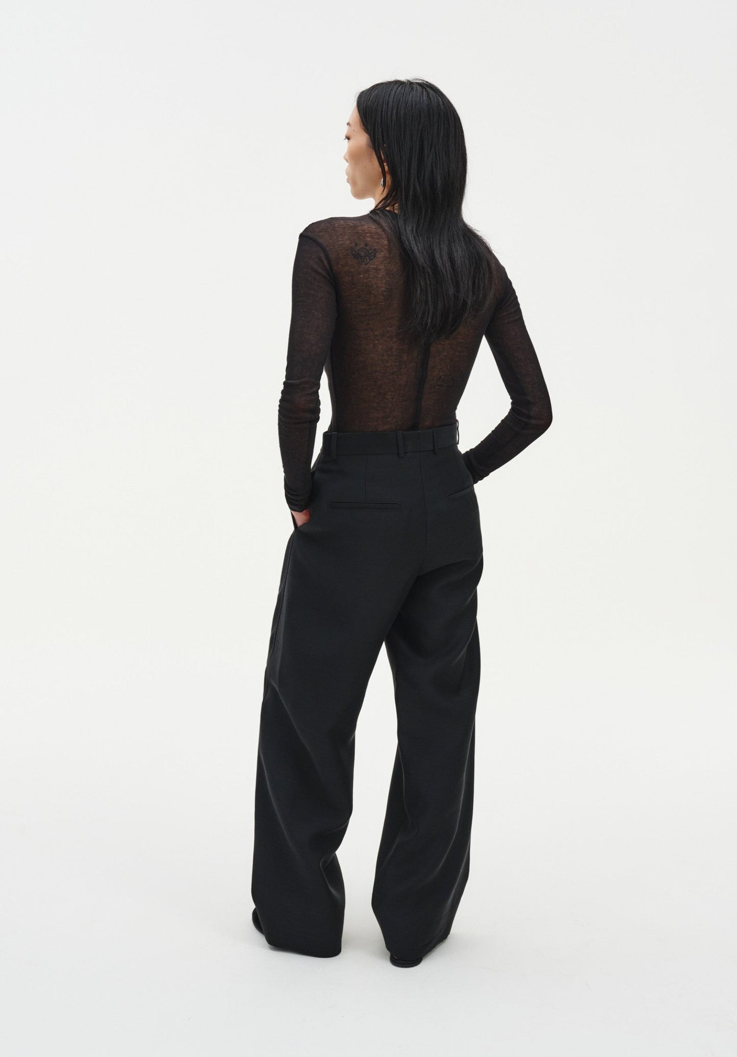 Rear three-quarter view of a woman against a plain white background, wearing a sheer long-sleeve dark top tucked into black tailored trousers. Her dark shoulder-length hair falls down her back, and one hand rests in her pocket, highlighting the relaxed fit and clean lines of the outfit.
