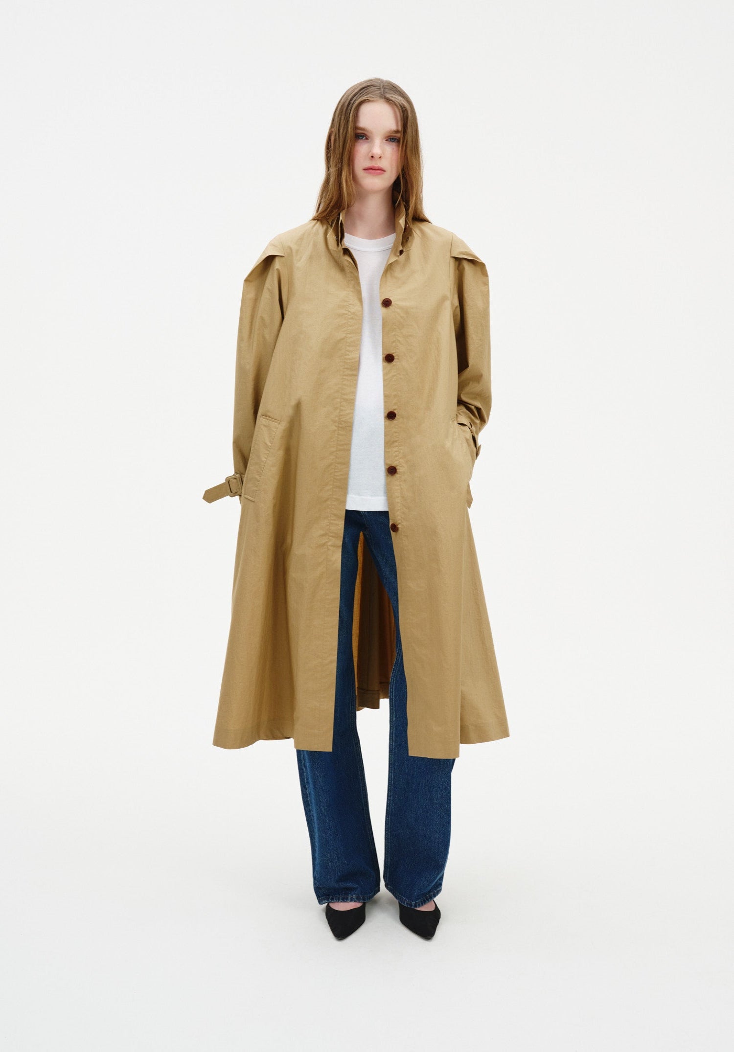 Front view of a model wearing a beige trench-style coat with button front and structured shoulders, layered over a white top and wide-leg blue jeans.
