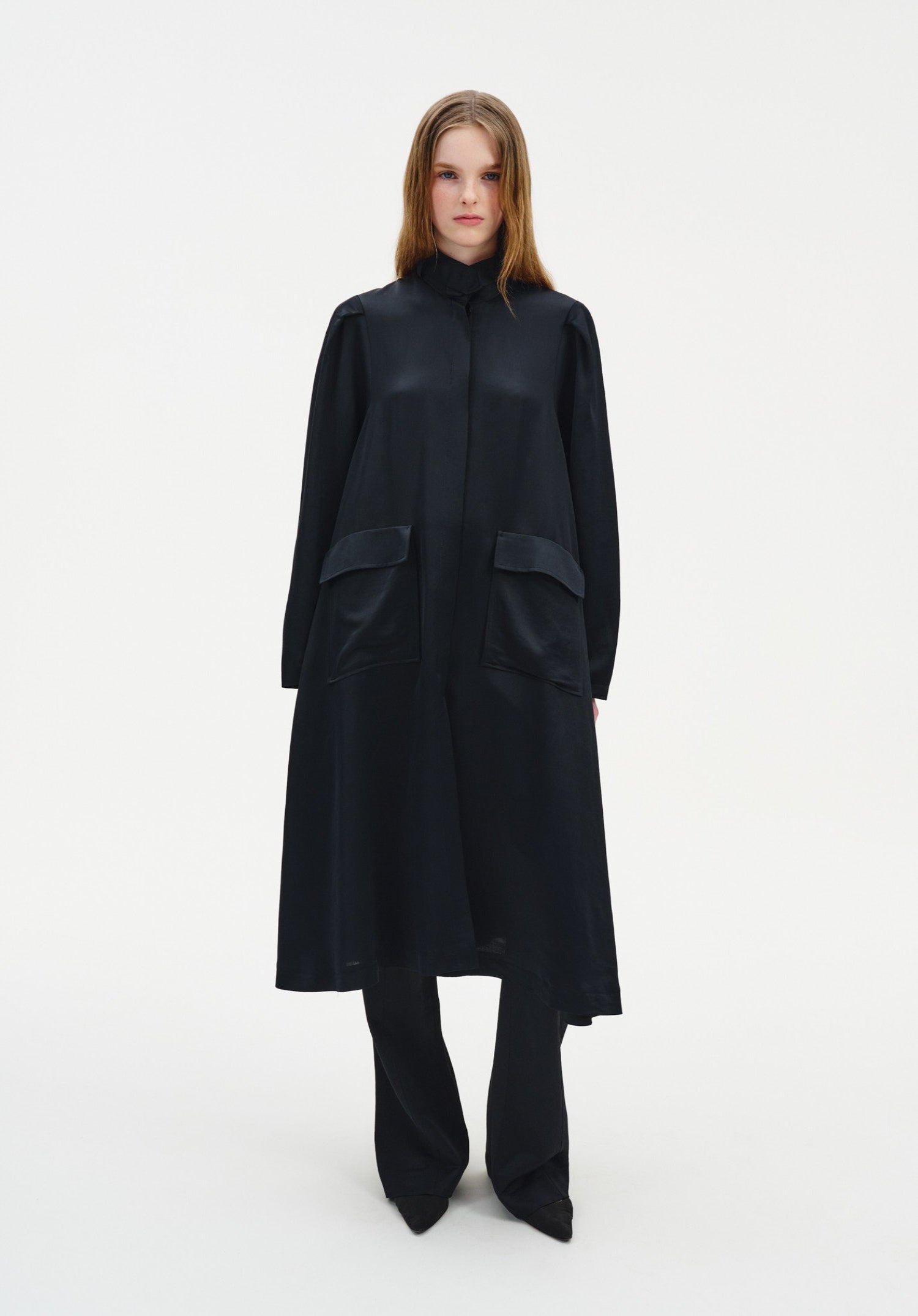 Front view of a model wearing a long, black, semi-sheer coat-style dress with a high neckline, long sleeves, and large patch pockets, layered over matching wide-leg trousers and paired with black pointed shoes.
