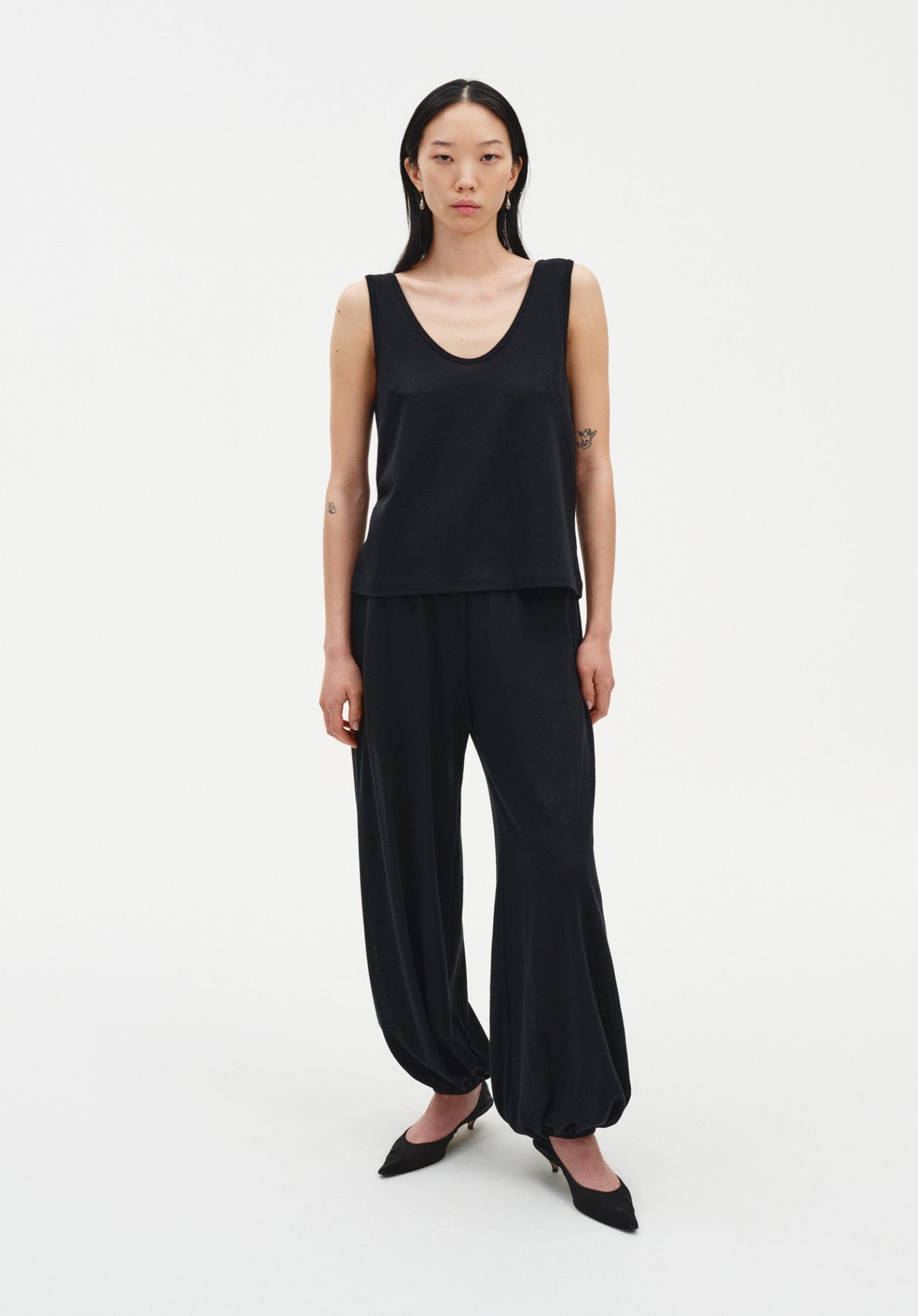 Front view of a woman wearing a black sleeveless scoop-neck tank top with a relaxed fit, paired with loose, draped black trousers gathered at the ankles and black pointed heels.
