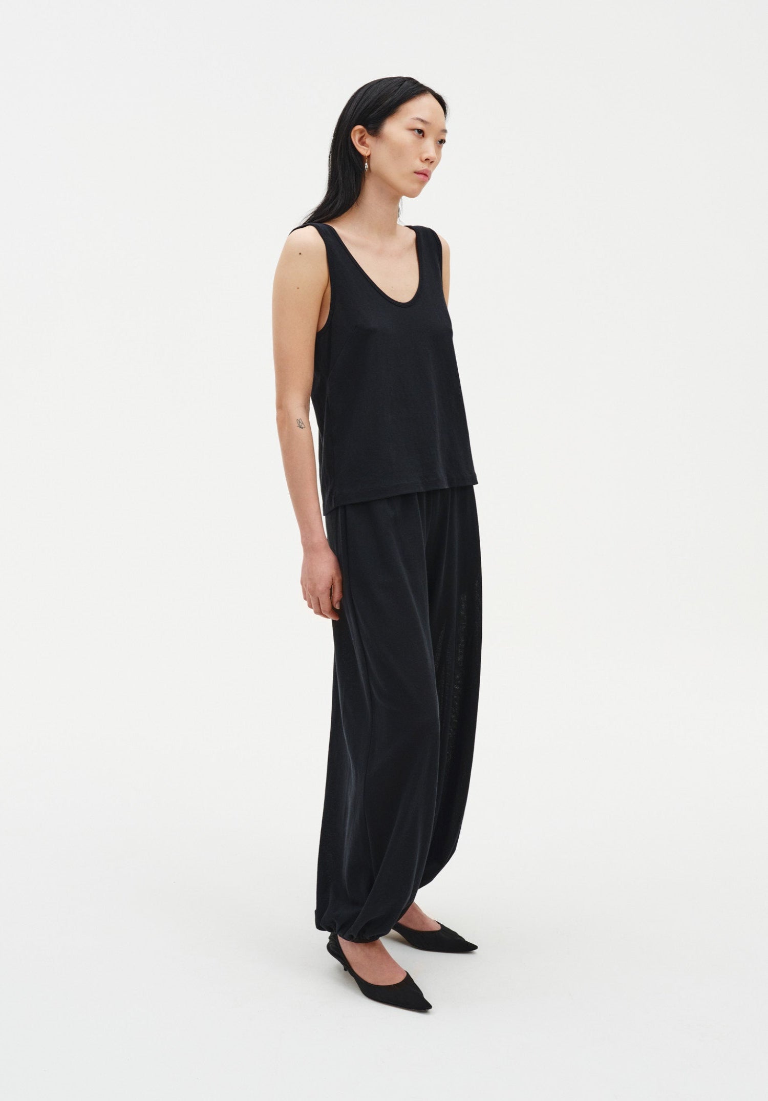 Three-quarter side view of a woman wearing a black sleeveless scoop-neck tank top with a relaxed silhouette, paired with loose, fluid black trousers gathered at the ankles and black pointed heels.
