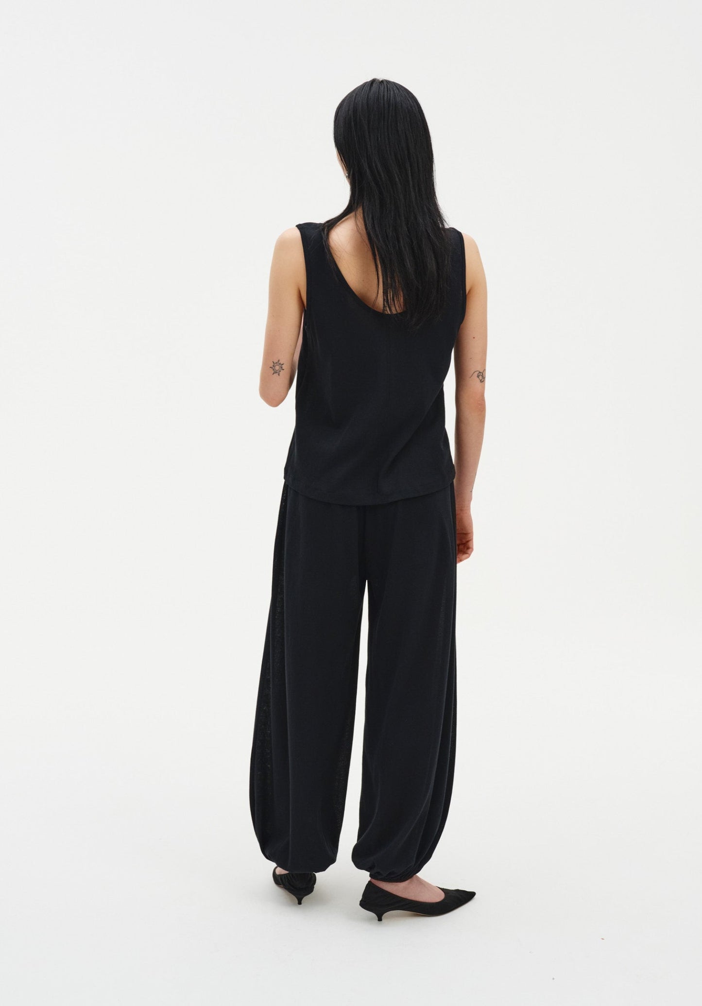 Back view of a woman wearing a black sleeveless tank top with a relaxed fit and scoop back, paired with loose, draped black trousers gathered at the ankles and black pointed heels.

