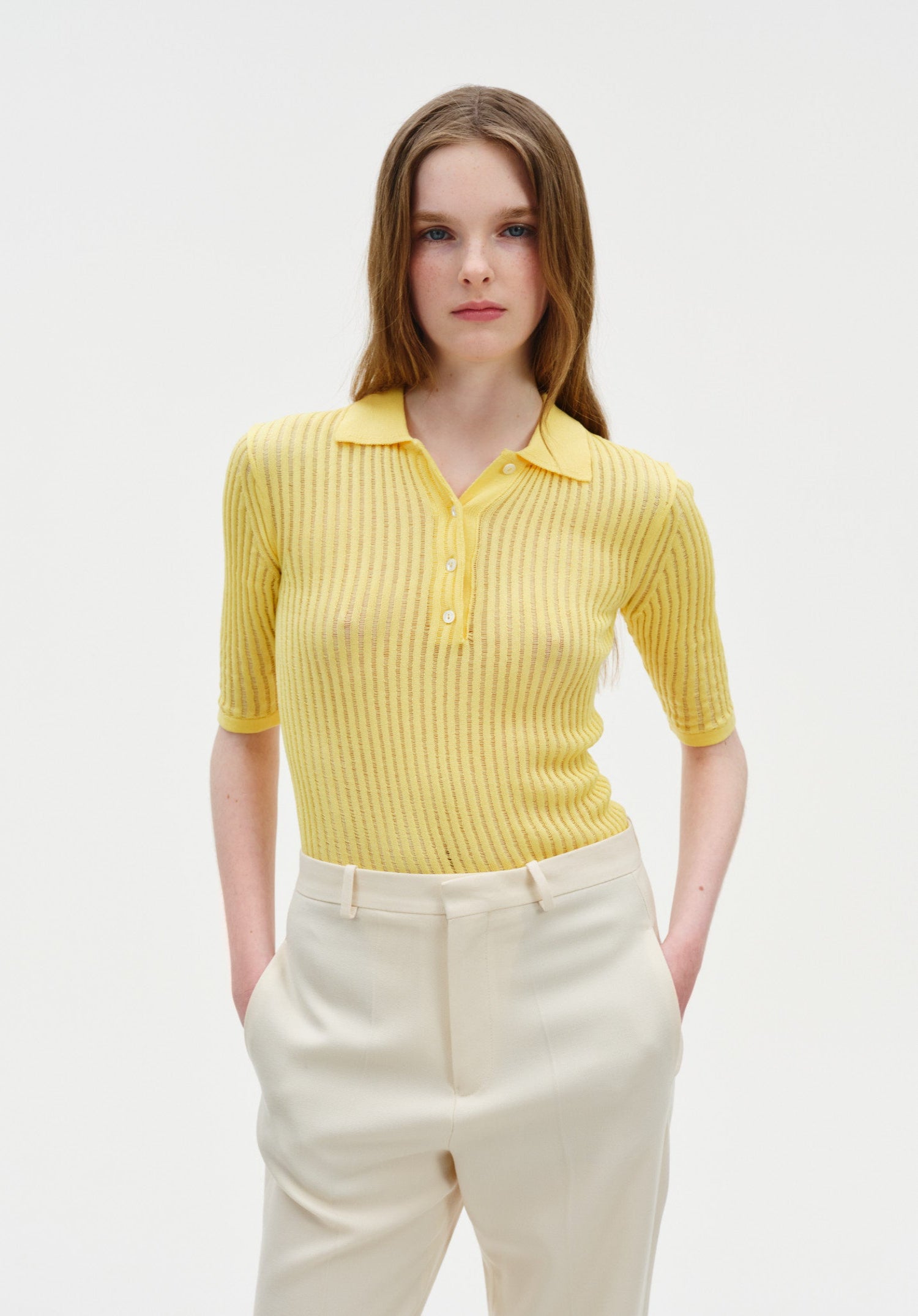 Front view of a model wearing a fitted yellow ribbed short-sleeve polo top with buttons, tucked into high-waisted cream trousers with hands in pockets.
