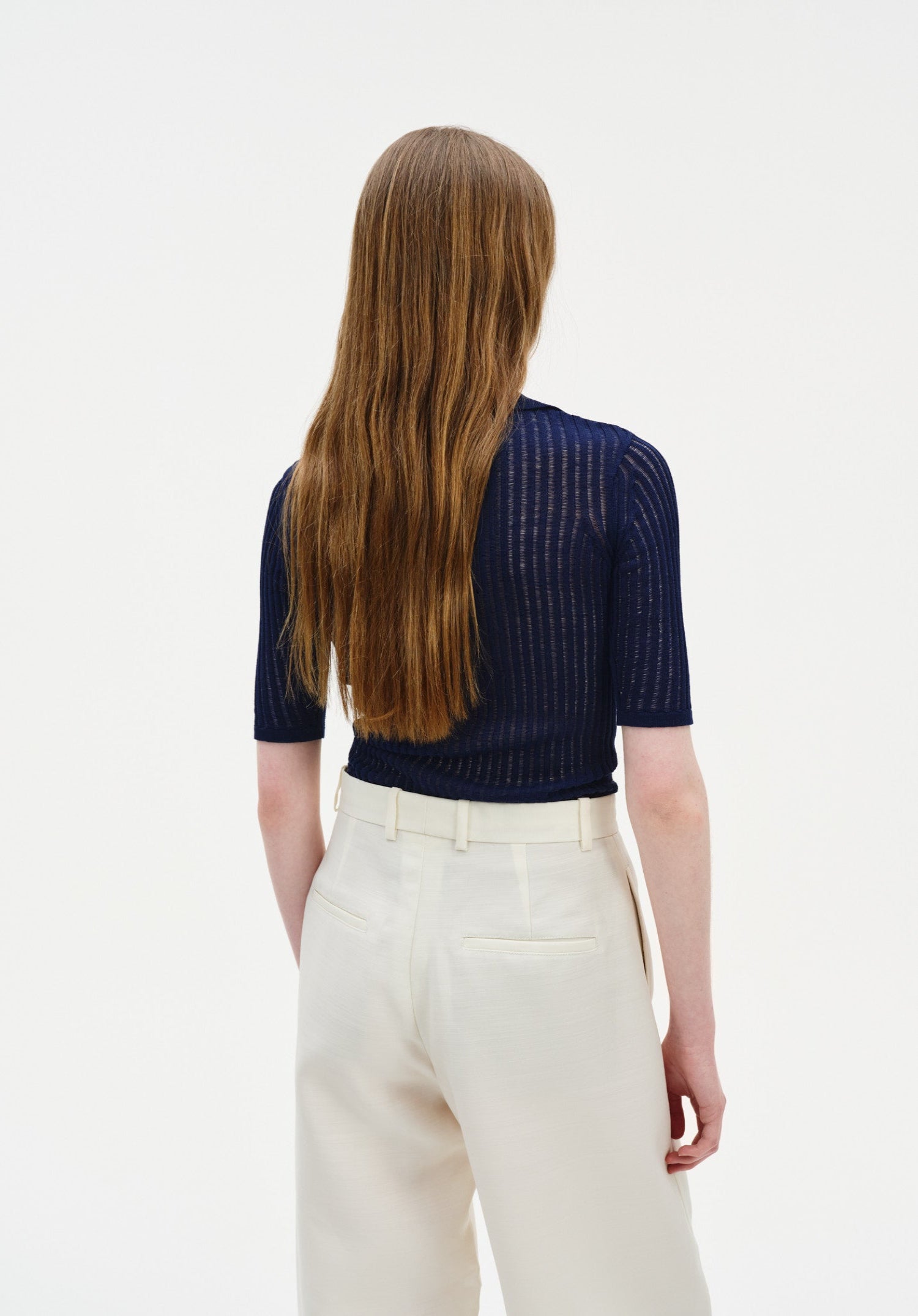 A woman with long light brown hair is shown from the back in a relaxed standing pose against a plain background, with her head slightly turned and her posture upright.
