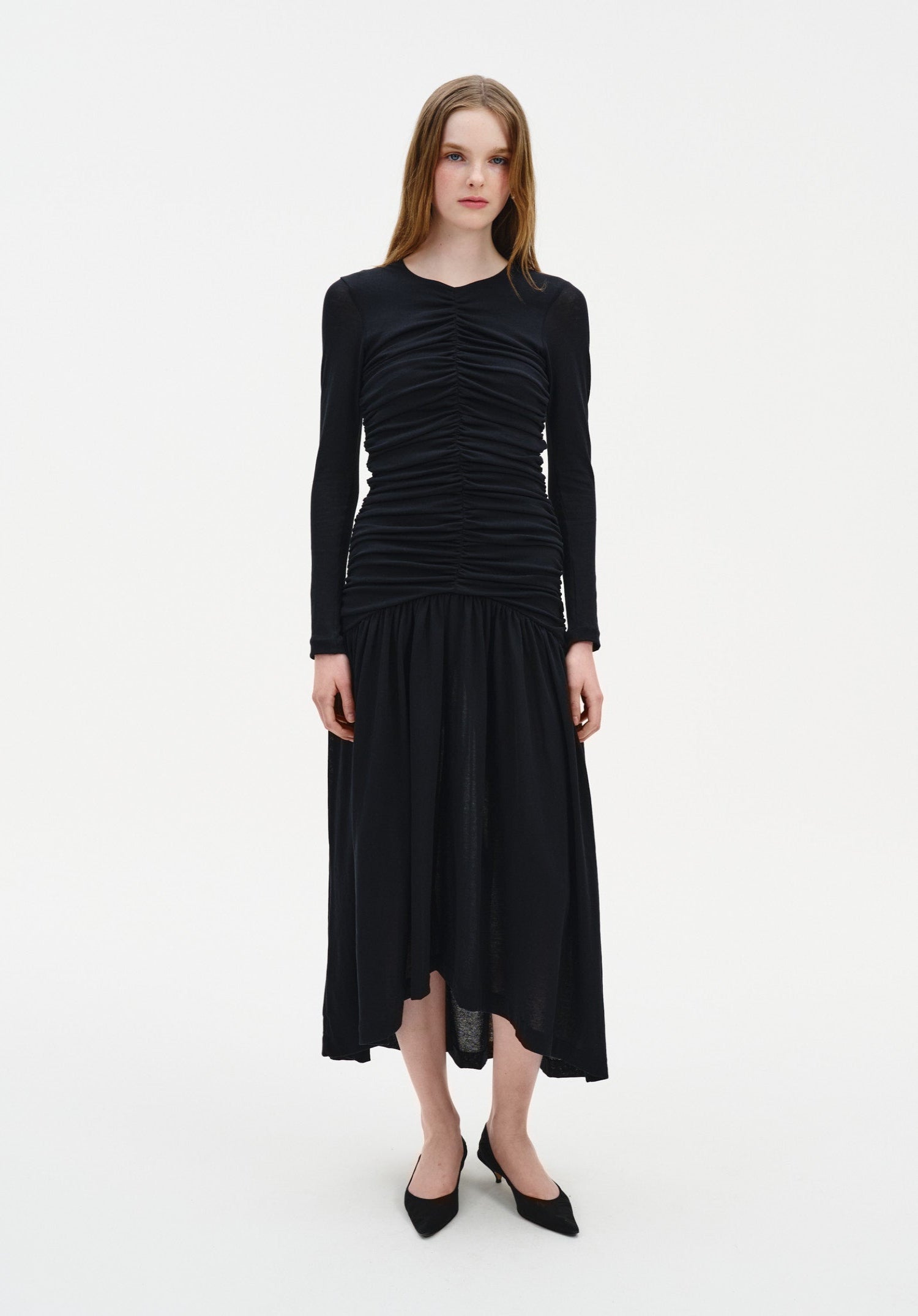 A woman stands facing forward against a plain white background, wearing a black long-sleeve dress with ruched detailing on the bodice and a loose, mid-calf skirt. She has straight, light brown hair worn down and is dressed with black pointed-toe heels, maintaining a neutral expression with arms relaxed at her sides.
