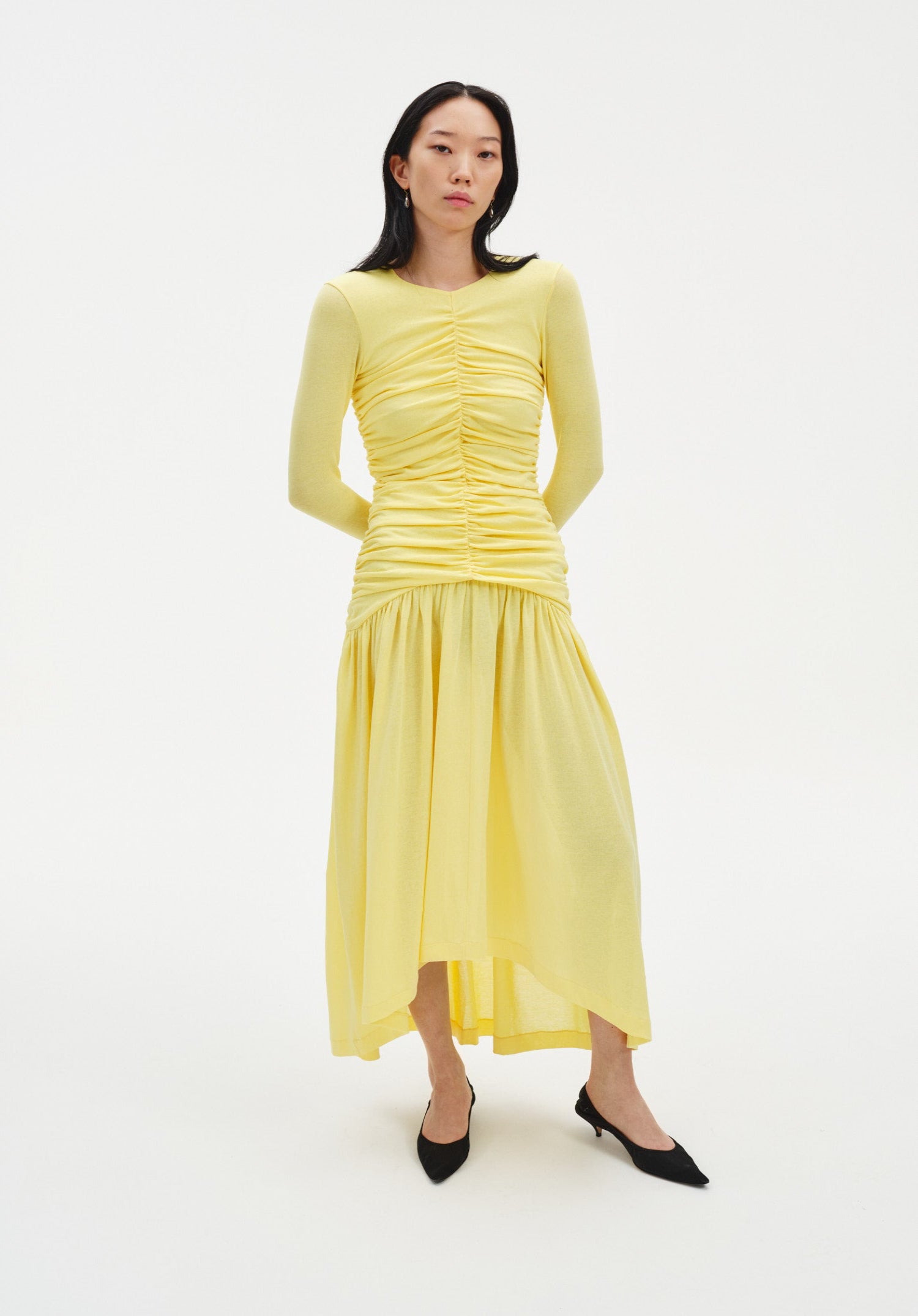Full-body front view of a woman standing with a relaxed posture and hands behind her back, facing forward with a neutral expression against a plain studio background.
