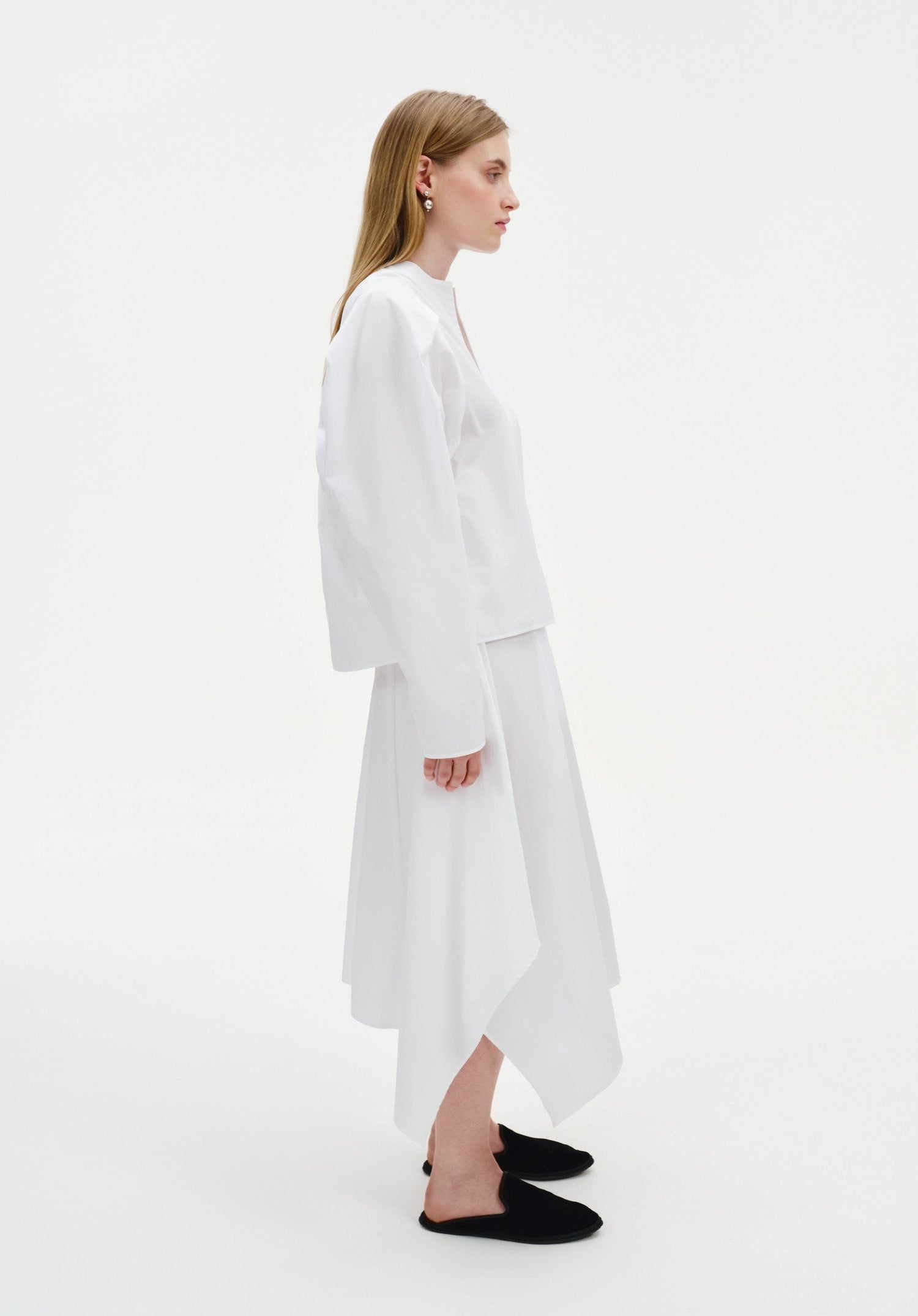 Side view of a woman wearing a white long-sleeve top with a clean, minimal silhouette paired with a white asymmetrical midi skirt, styled with black slip-on flats.
