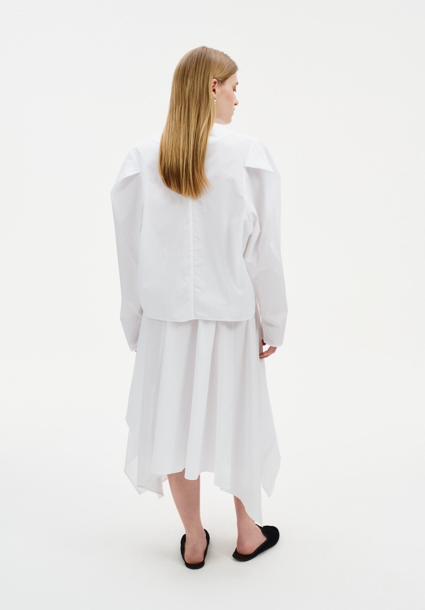 Back view of a woman wearing a white long-sleeve top with structured shoulders and a clean, minimal cut, paired with a flowing white asymmetrical midi skirt and black slip-on flats.
