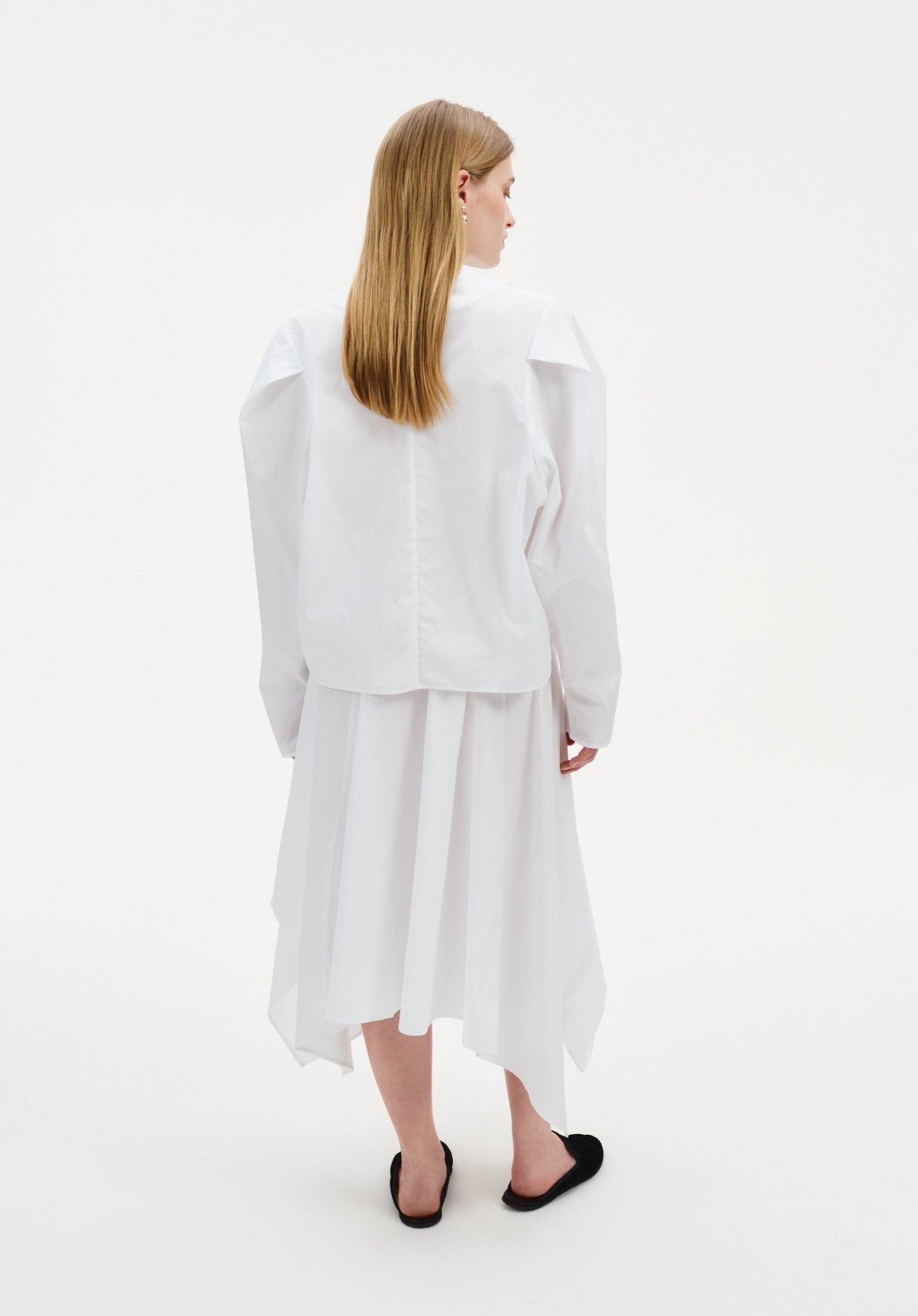 Back view of a woman wearing a white long-sleeve top with structured shoulders and a clean, minimal cut, paired with a flowing white asymmetrical midi skirt and black slip-on flats.
