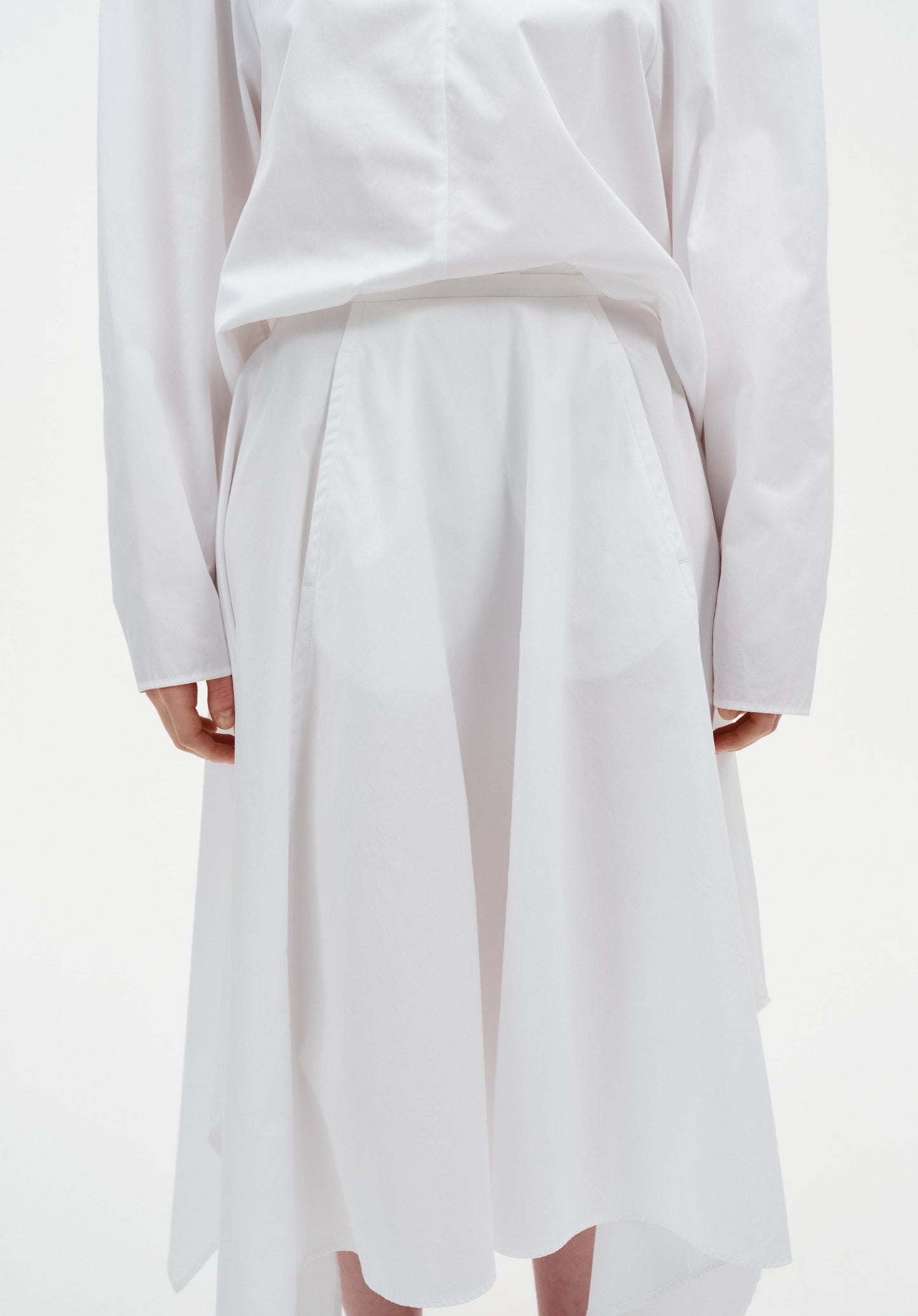 Close-up of a white outfit showing a long-sleeve top tucked into a high-waisted skirt with soft pleats and a fluid, asymmetrical hem.
