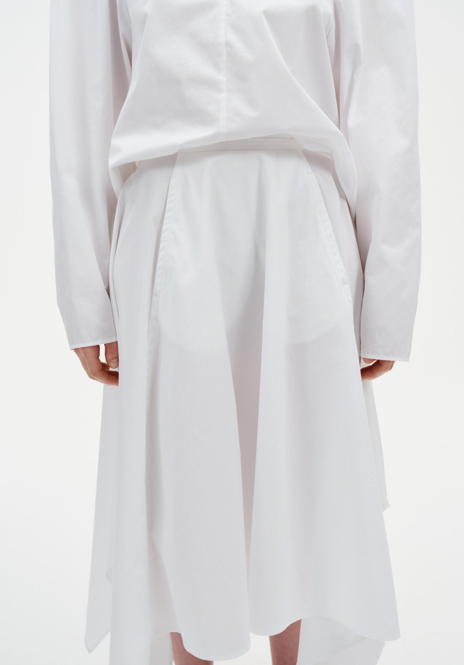 Close-up of a white outfit showing a long-sleeve top tucked into a high-waisted skirt with soft pleats and a fluid, asymmetrical hem.

