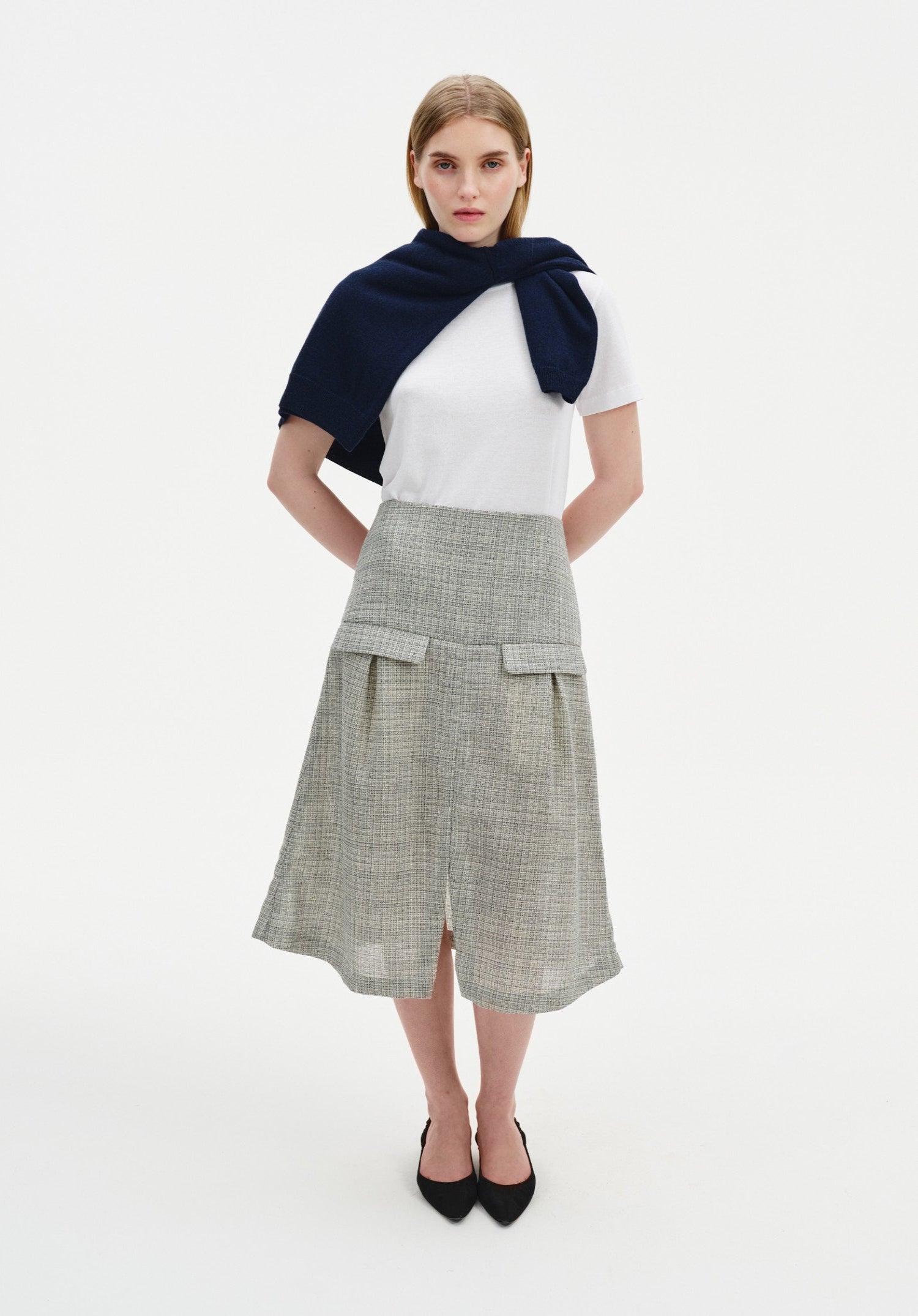 A woman stands facing forward with a slight three-quarter angle against a plain white background, wearing a white short-sleeve top with a navy sweater draped over her shoulders, paired with a light gray textured midi skirt with flap pockets and a front slit, and black pointed heels.
