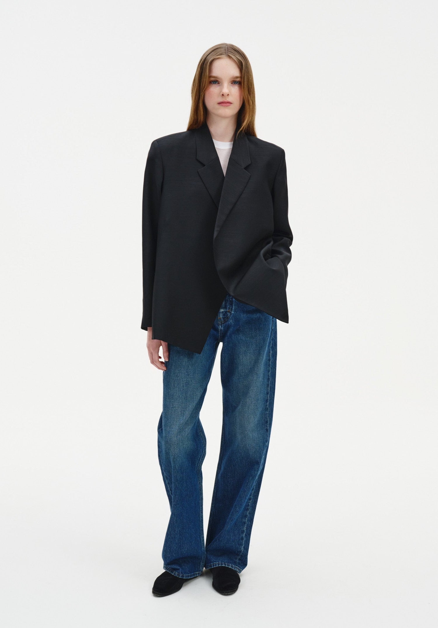 A woman with long light brown hair stands facing forward against a plain background, posing in a relaxed stance with one hand in her pocket and a neutral, composed expression.
