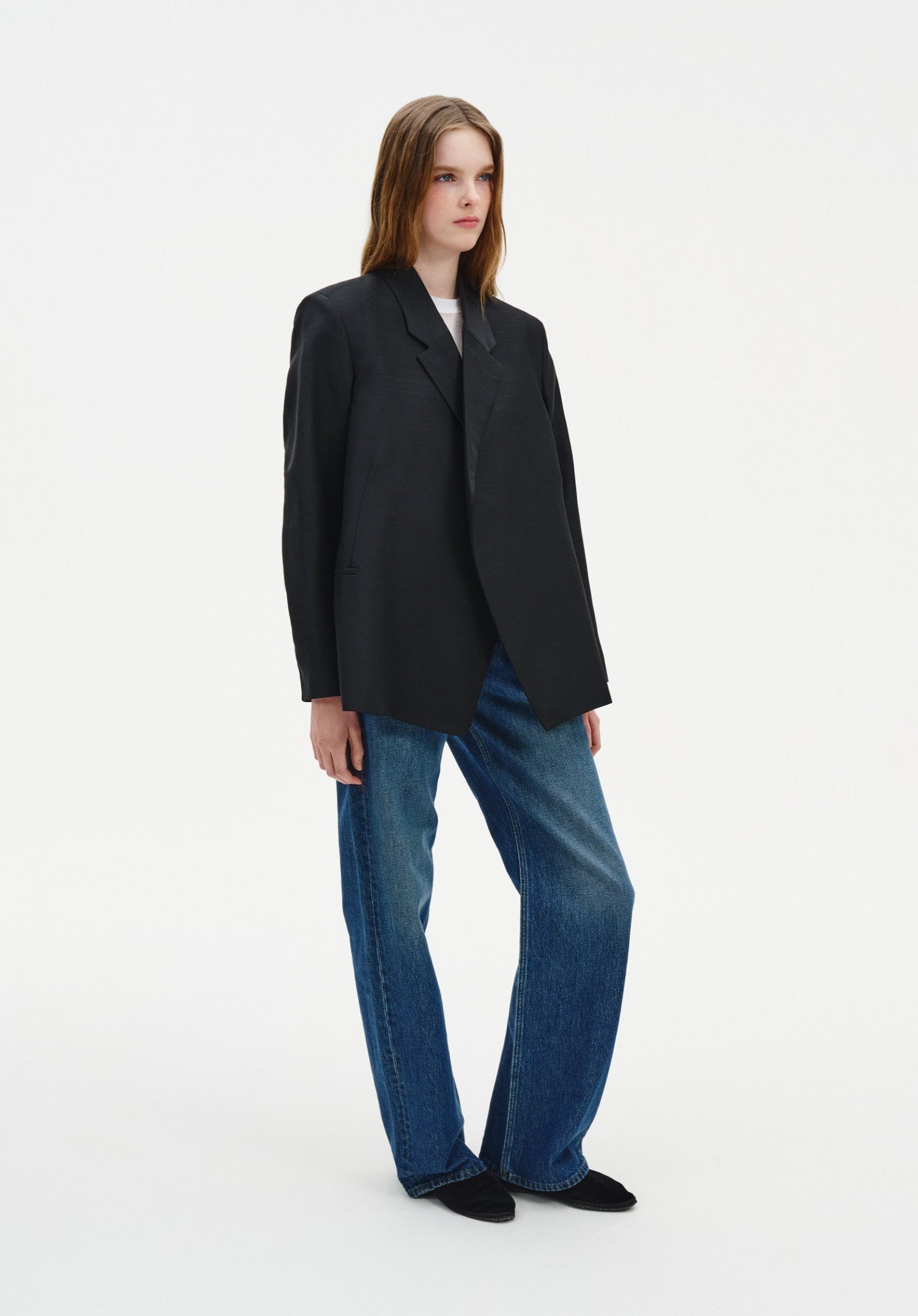 A woman with long light brown hair stands in a relaxed three-quarter pose against a plain background, looking slightly to the side with a calm, neutral expression.
