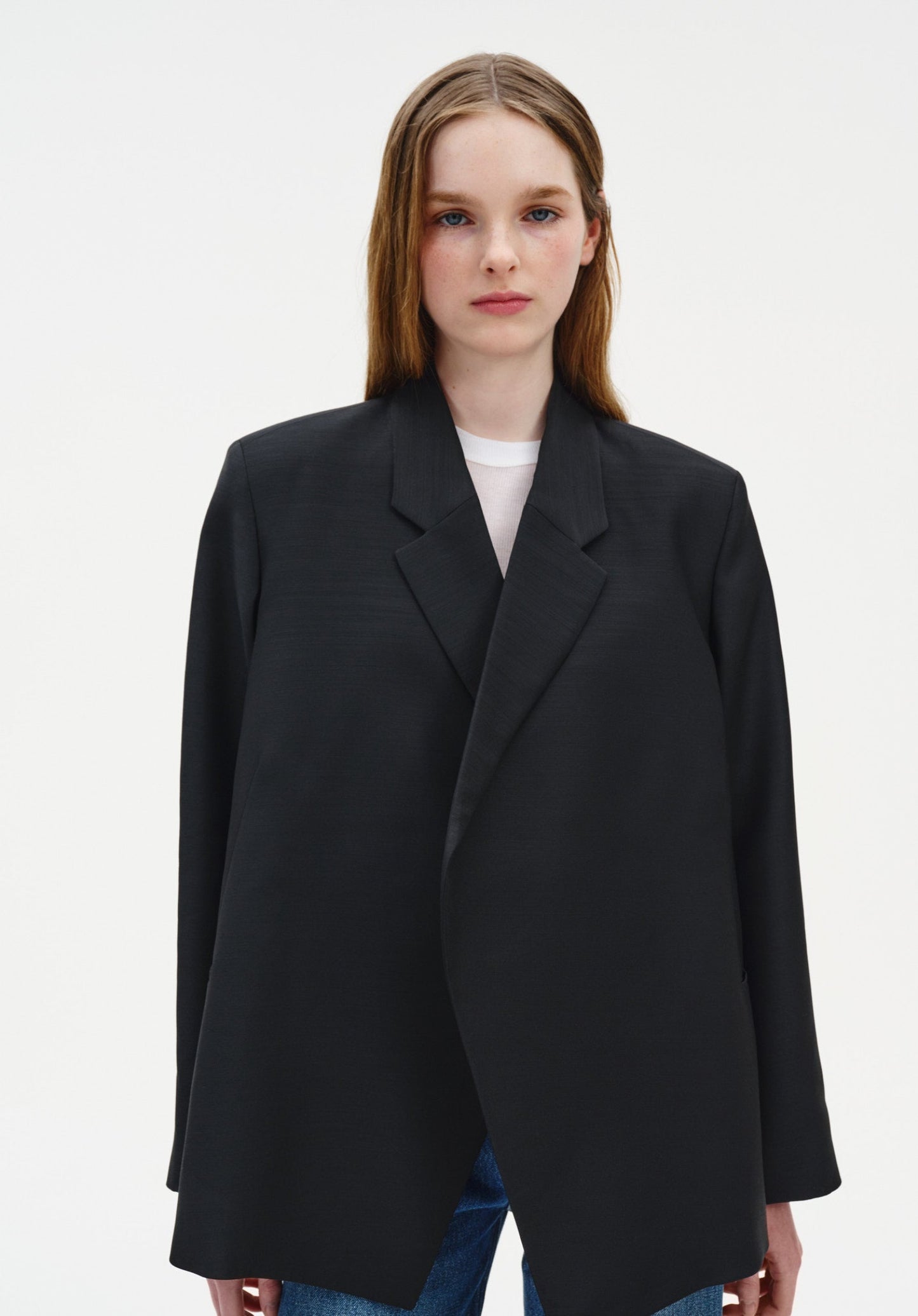 A woman with straight light brown hair stands facing forward against a plain background, maintaining a neutral expression with a relaxed, upright posture and her gaze directed at the camera.
