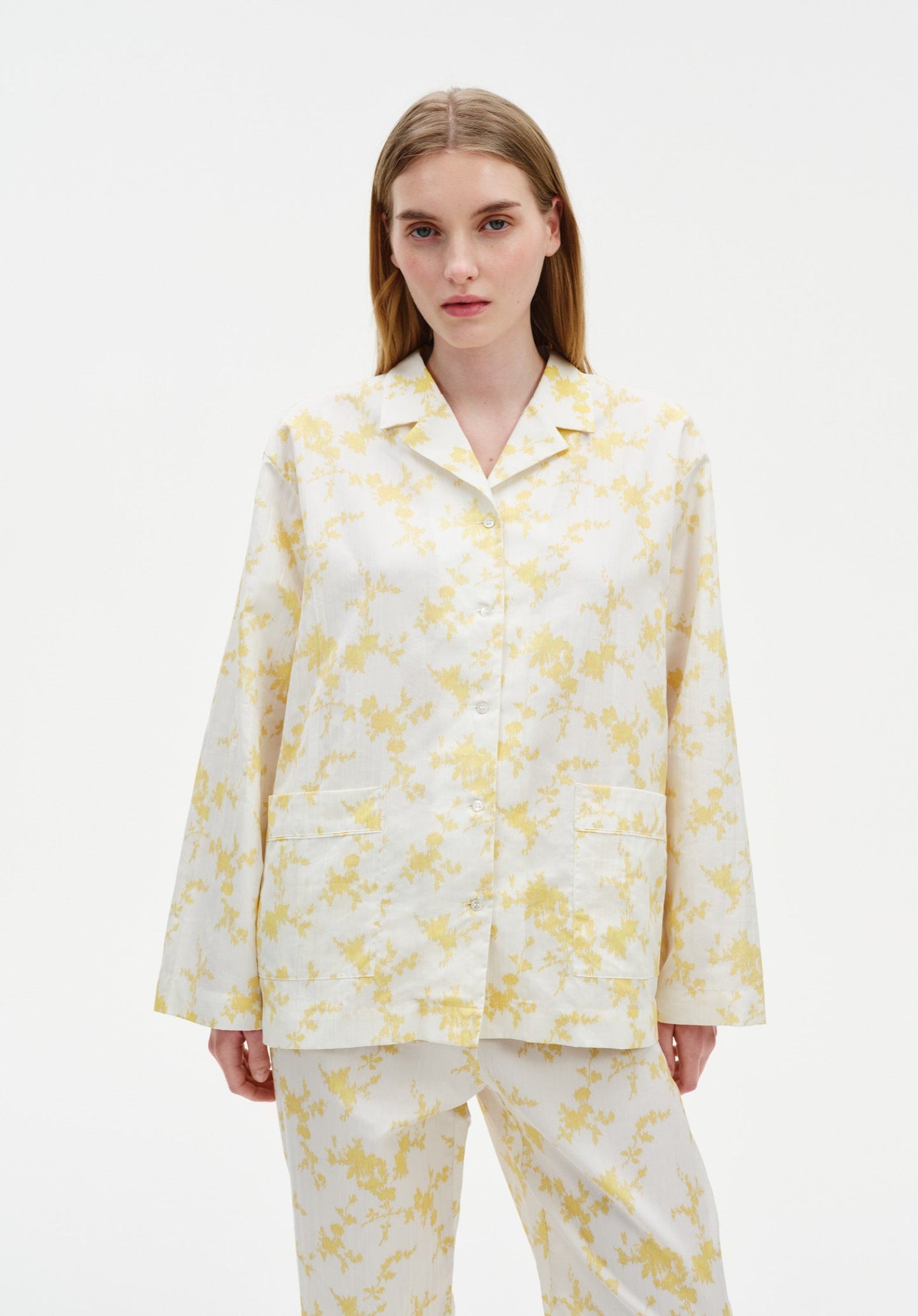 Front view of a woman wearing a white button-up shirt and matching trousers with a soft yellow floral pattern, featuring a relaxed fit with a notched collar and patch pockets, set against a plain studio background.
