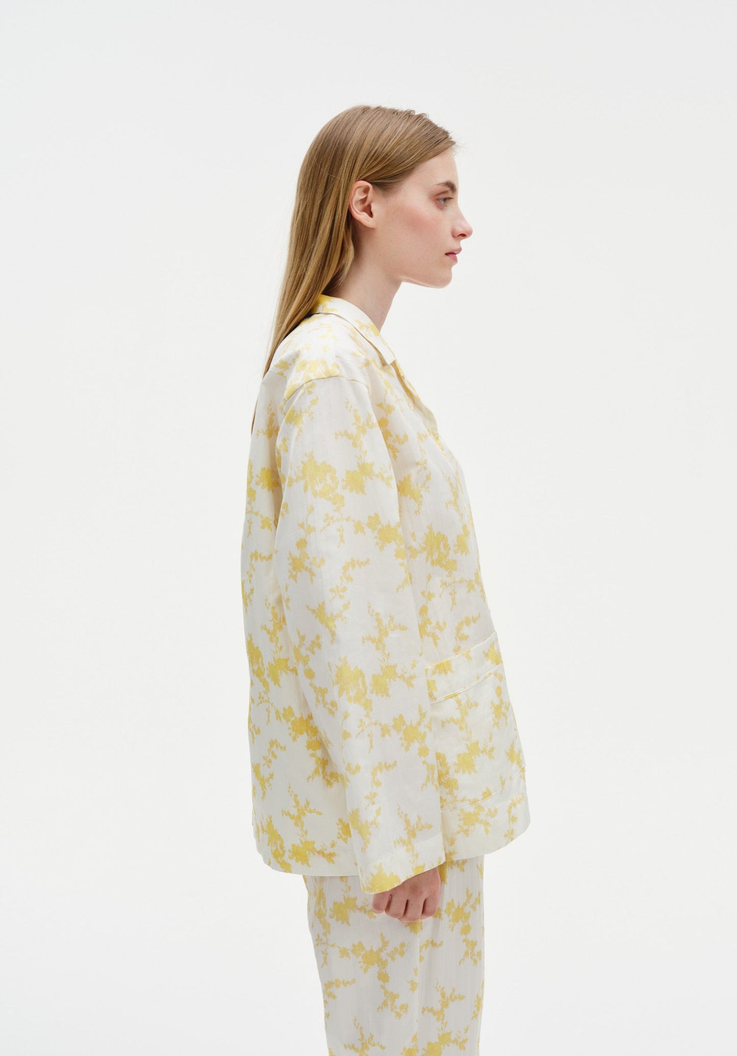 Side profile of a woman wearing a white long-sleeve shirt and matching trousers with a soft yellow floral pattern, highlighting the relaxed fit and clean silhouette against a plain studio background.
