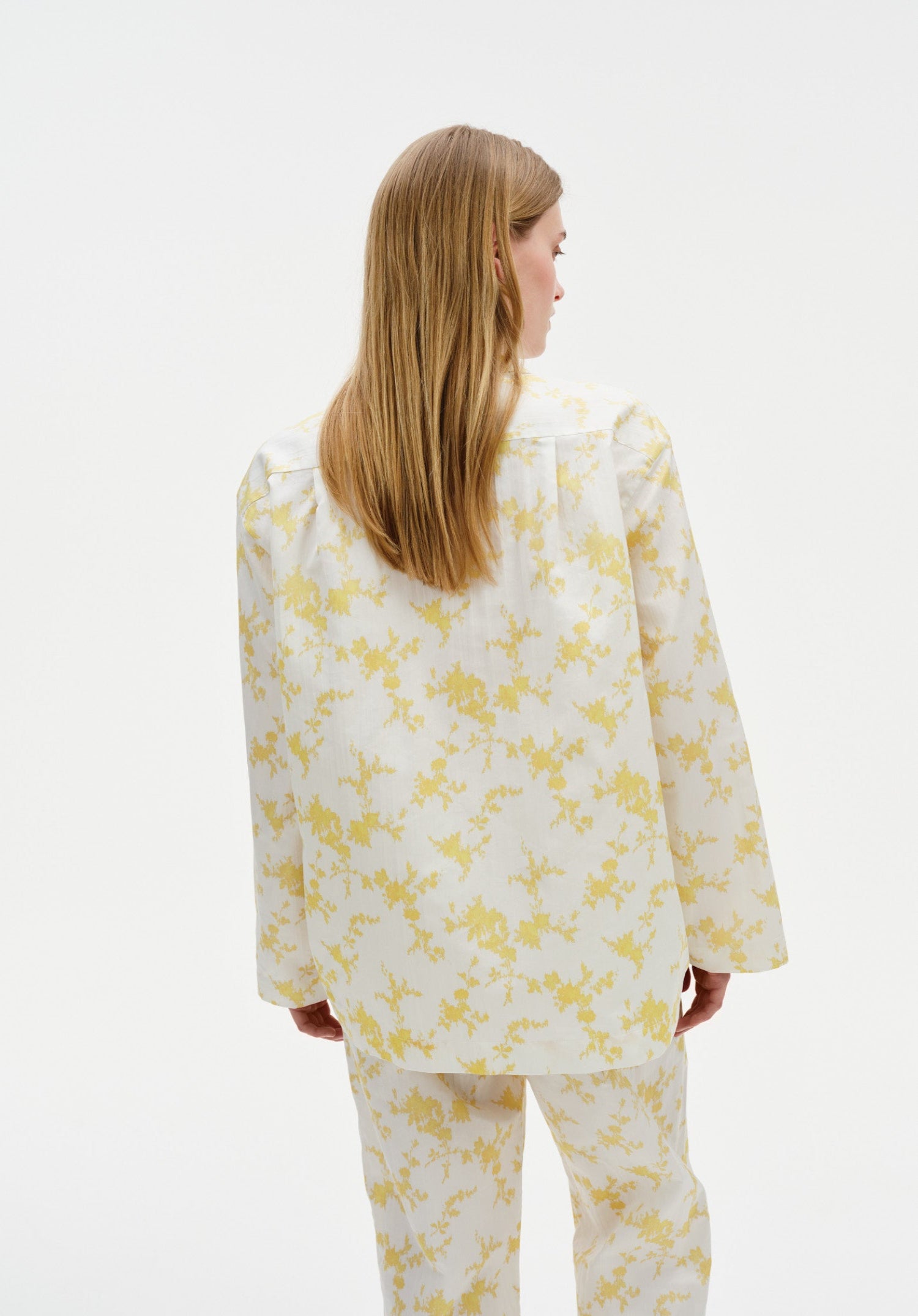 Back view of a woman wearing a white long-sleeve top and matching trousers with a soft yellow floral pattern, showing the relaxed fit and drape of the outfit against a plain studio background.
