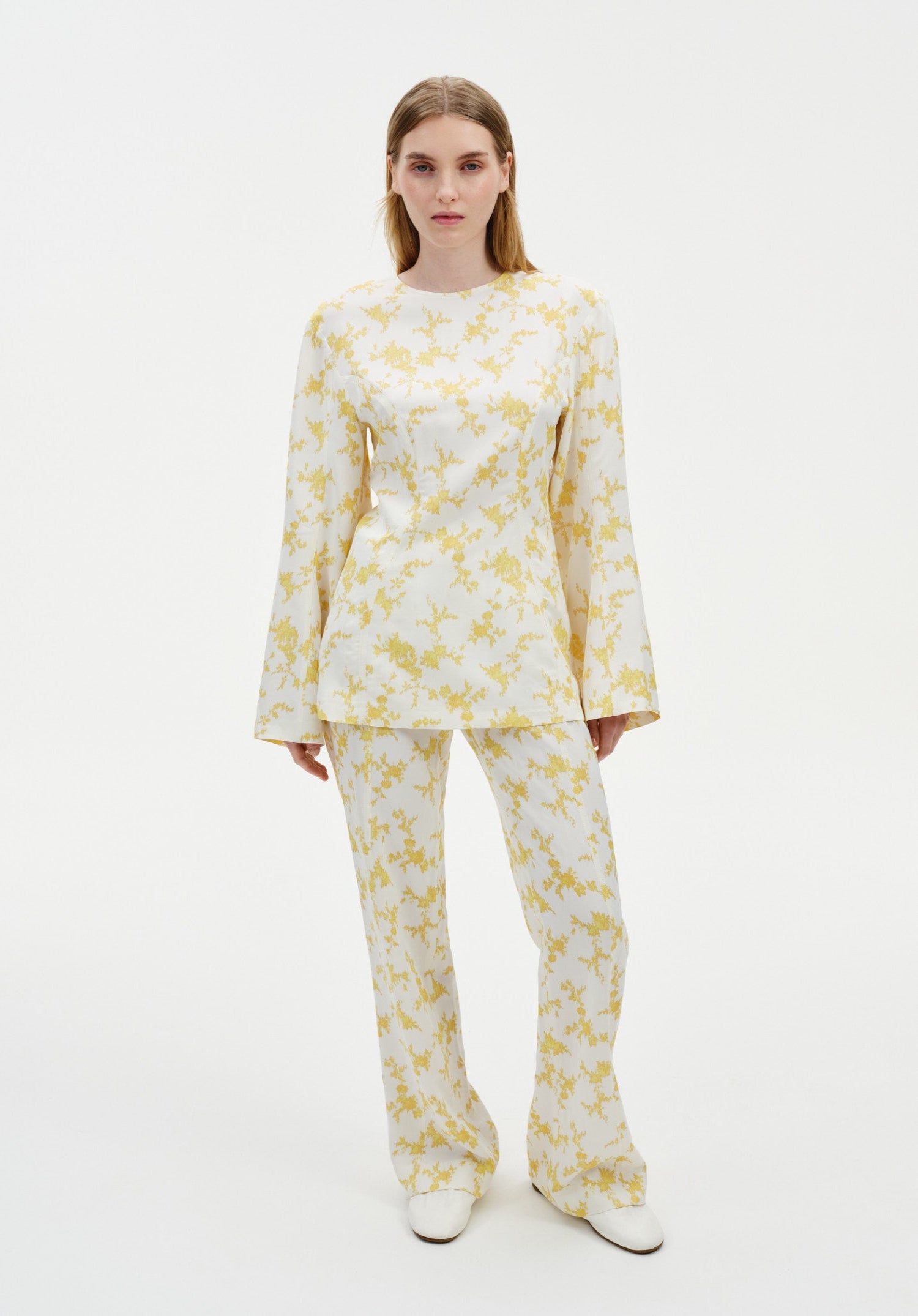 Model wearing a coordinated cream outfit with a yellow abstract floral print, featuring a long-sleeve tunic top with flared cuffs and matching straight-leg trousers, styled with white shoes.
