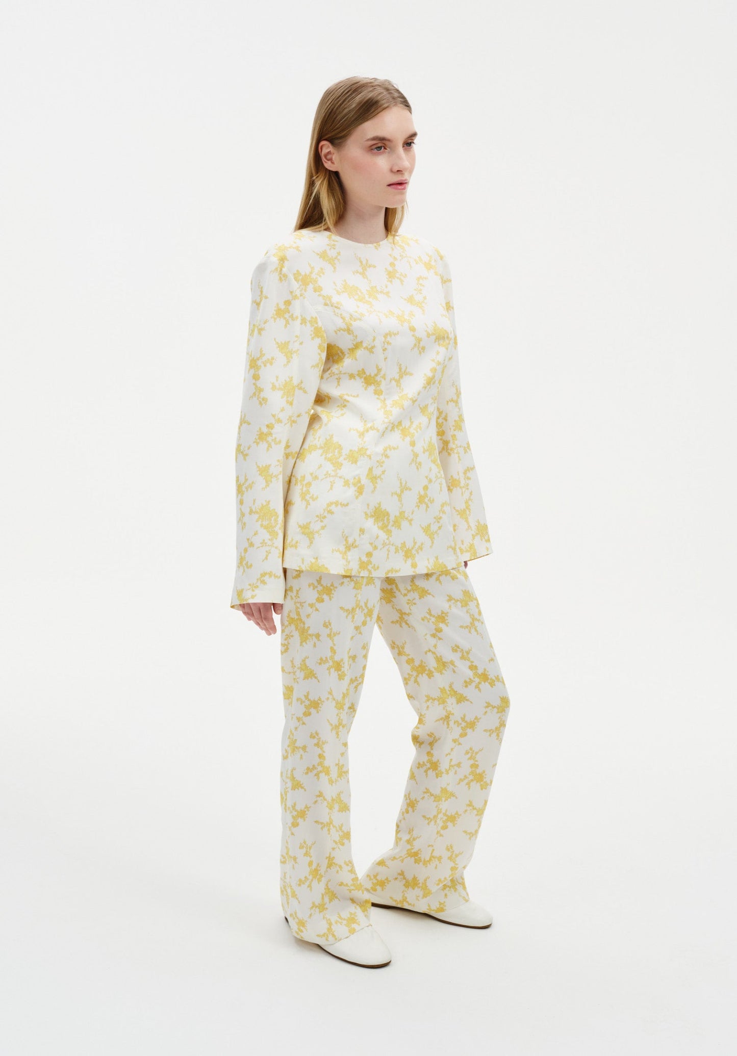 Back view of a model wearing a cream two-piece outfit with a yellow abstract floral print, featuring a long-sleeve tunic top and matching straight-leg trousers.
