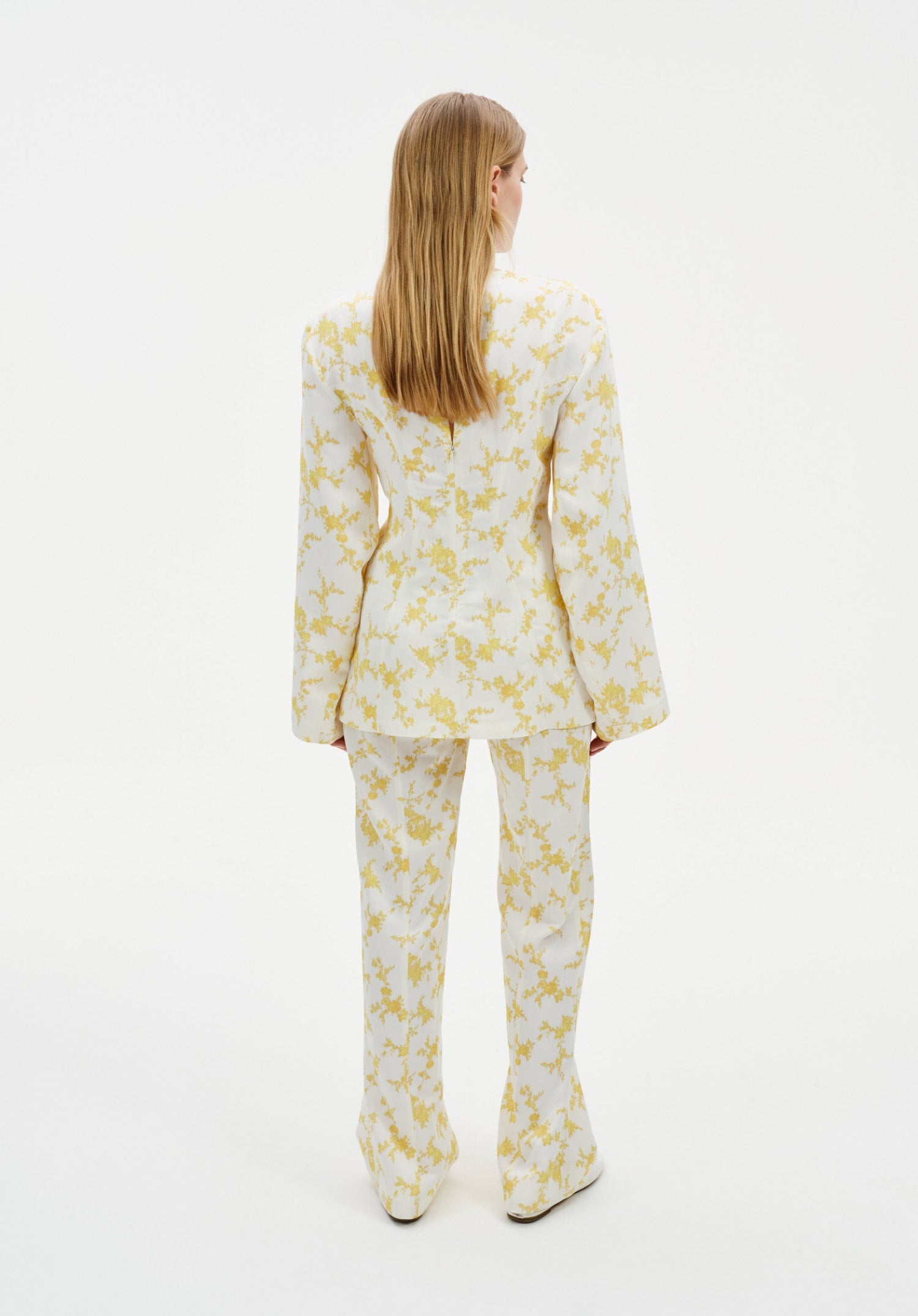 Back view of a model wearing a cream two-piece outfit with a yellow abstract floral print, featuring a long-sleeve tunic top and matching straight-leg trousers.
