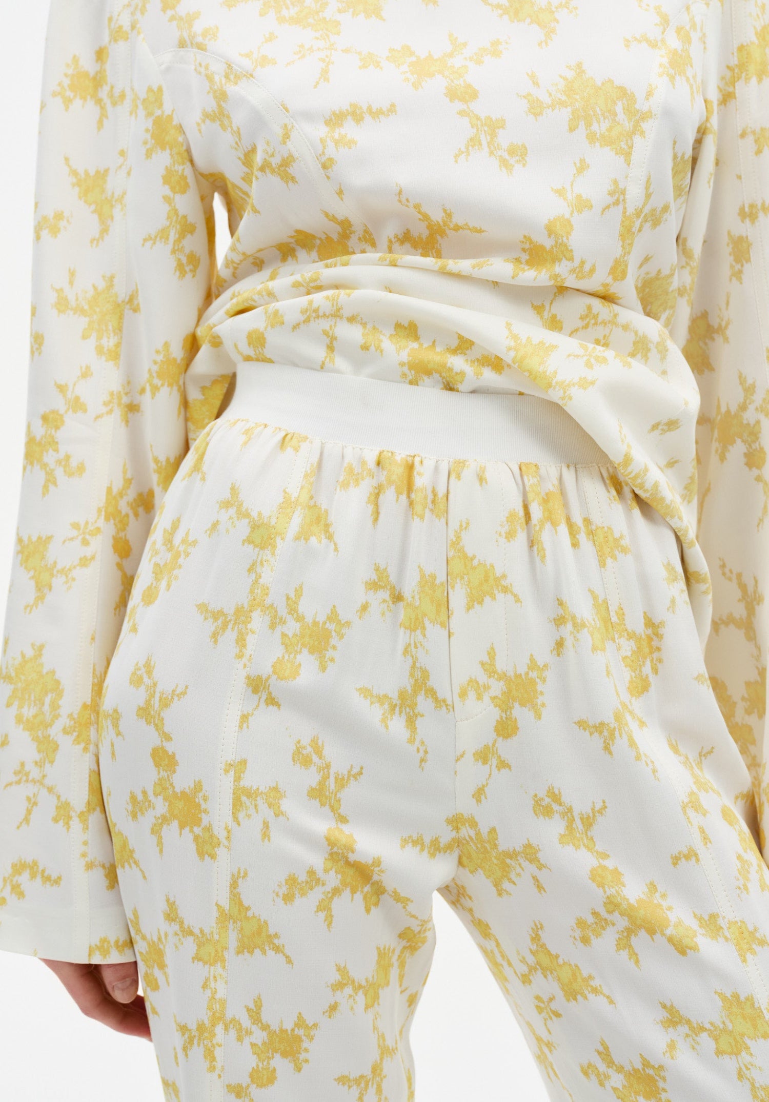 Close-up of cream trousers with a yellow abstract floral print, featuring a high elasticated waistband and soft draping fabric, paired with a matching long-sleeve top.

