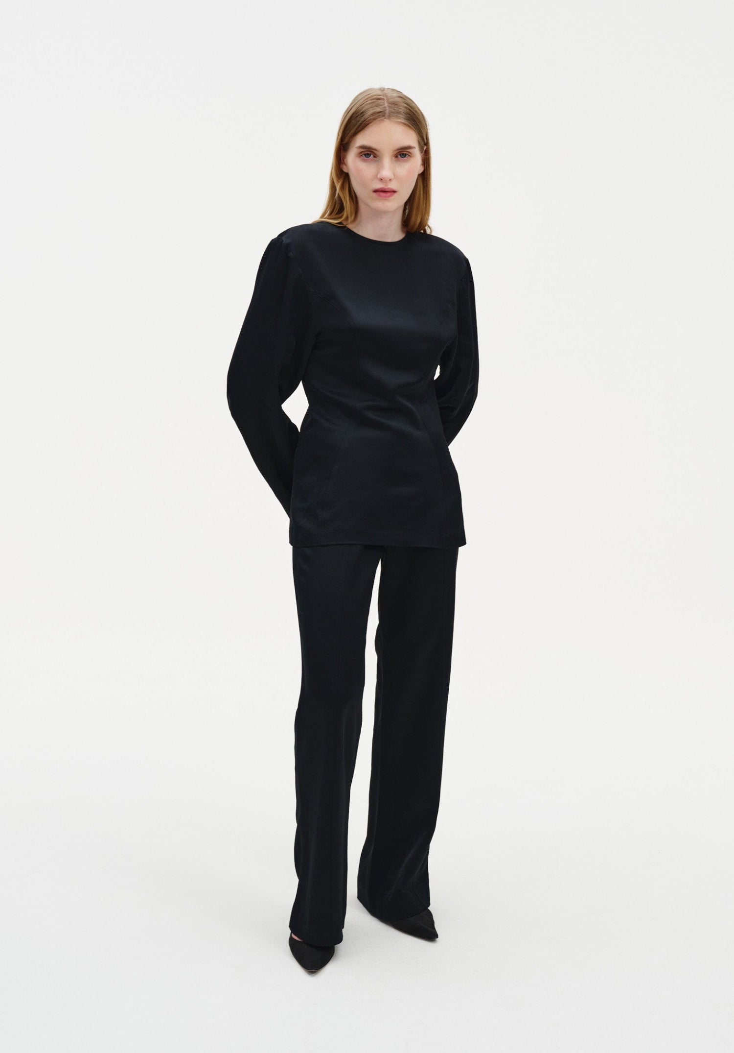 Full-length front view of a woman wearing a black long-sleeve top and matching tailored trousers, showcasing a sleek, fitted silhouette with a smooth finish against a plain studio background.
