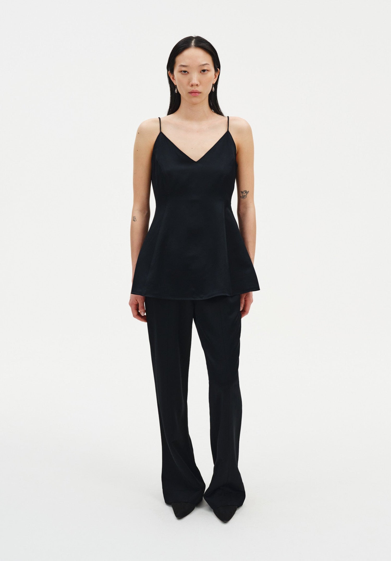A woman stands facing forward against a plain white background, wearing a black sleeveless top with thin straps and a flared hem, paired with black trousers and pointed shoes. Her dark shoulder-length hair is worn down, and small tattoos are visible on her arms as she maintains a neutral expression.
