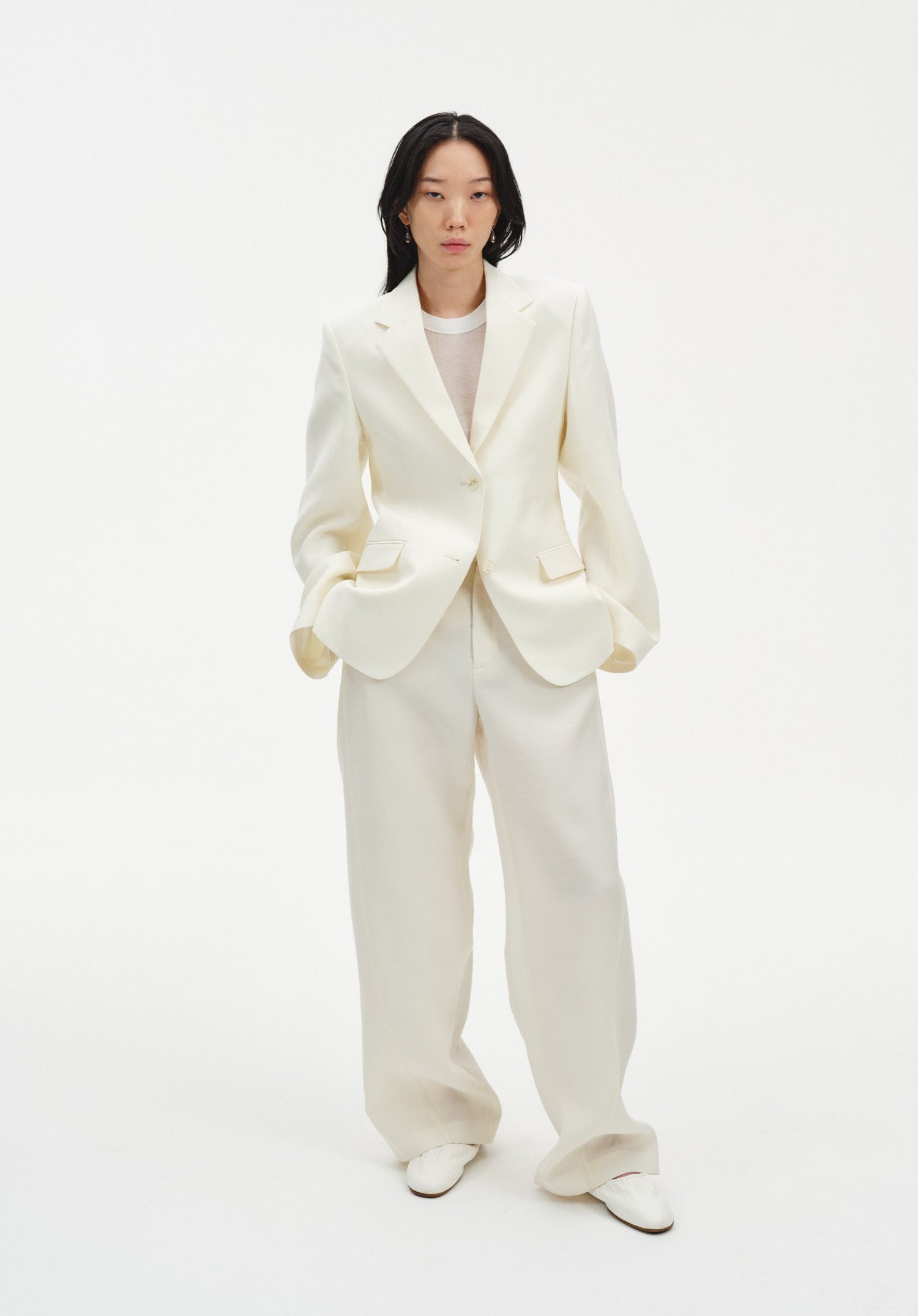 A woman stands facing forward against a plain white background, wearing a cream-colored tailored blazer with a single-button closure and matching wide-leg trousers, paired with light-colored shoes. Her dark shoulder-length hair is worn down, and she maintains a neutral expression with her hands in her pockets.
