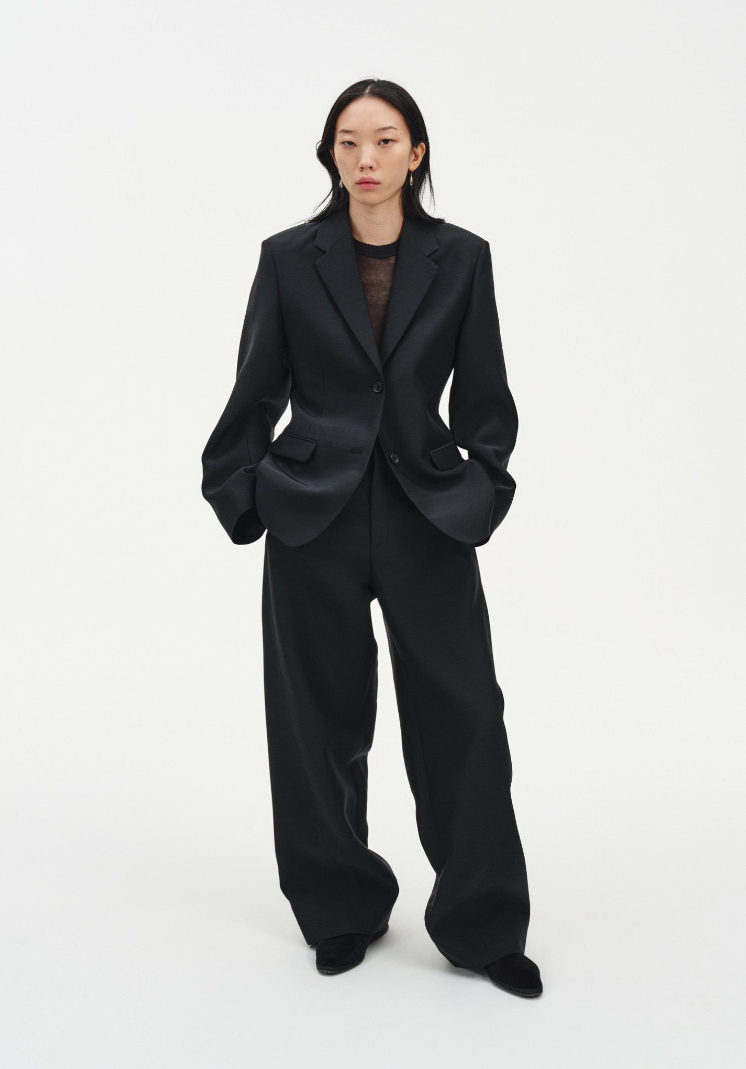 A woman stands facing forward against a plain white background, wearing a black tailored blazer over a sheer dark top with matching wide-leg trousers and black shoes. Her dark shoulder-length hair is worn down, and she maintains a neutral expression with her hands in her pockets.
