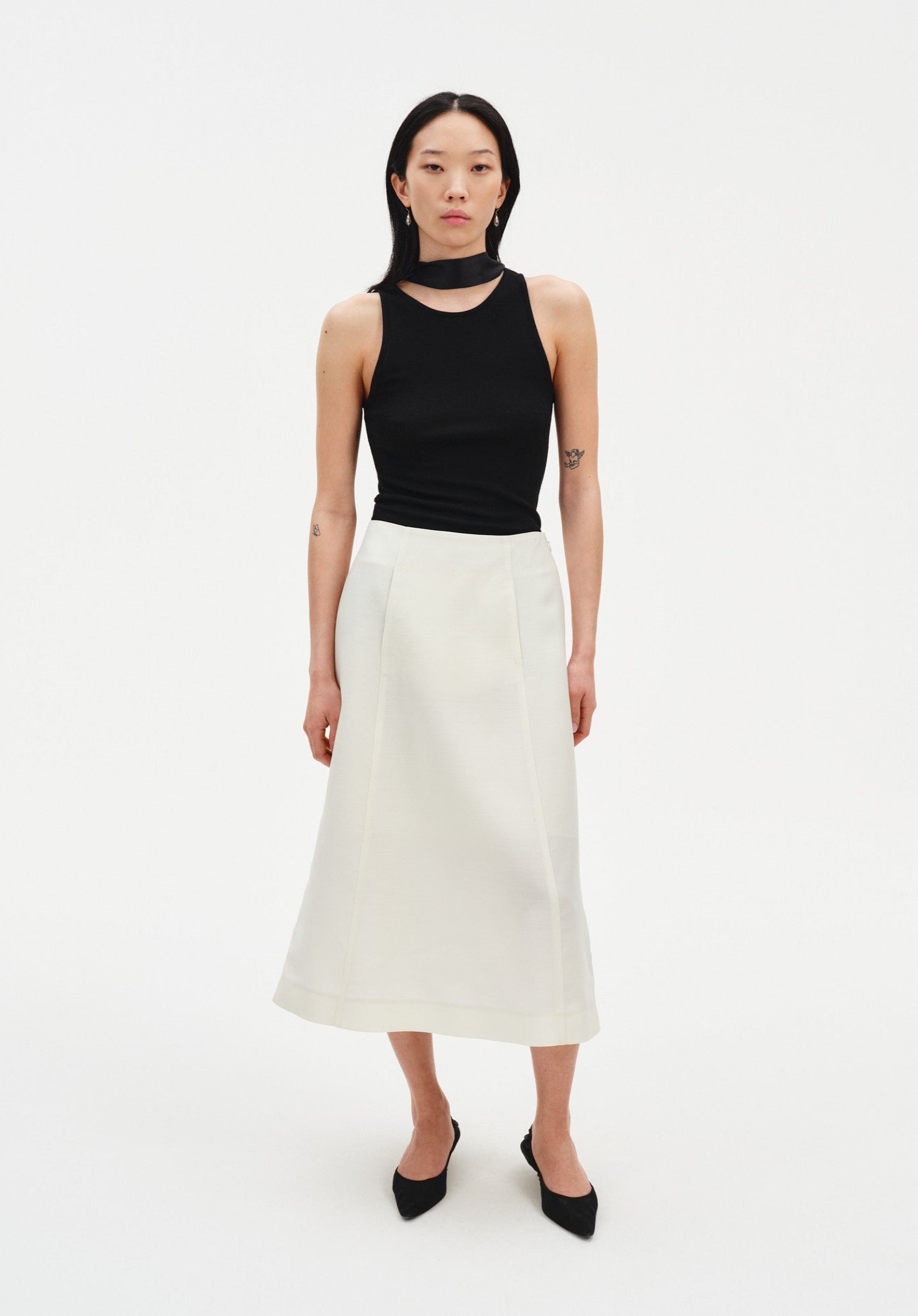 A woman stands facing forward against a plain white background, wearing a black sleeveless top with a high neckline and choker-style detail, paired with a cream-colored midi skirt and black pointed heels. Her dark shoulder-length hair is worn down, and she maintains a neutral expression with arms relaxed at her sides.
