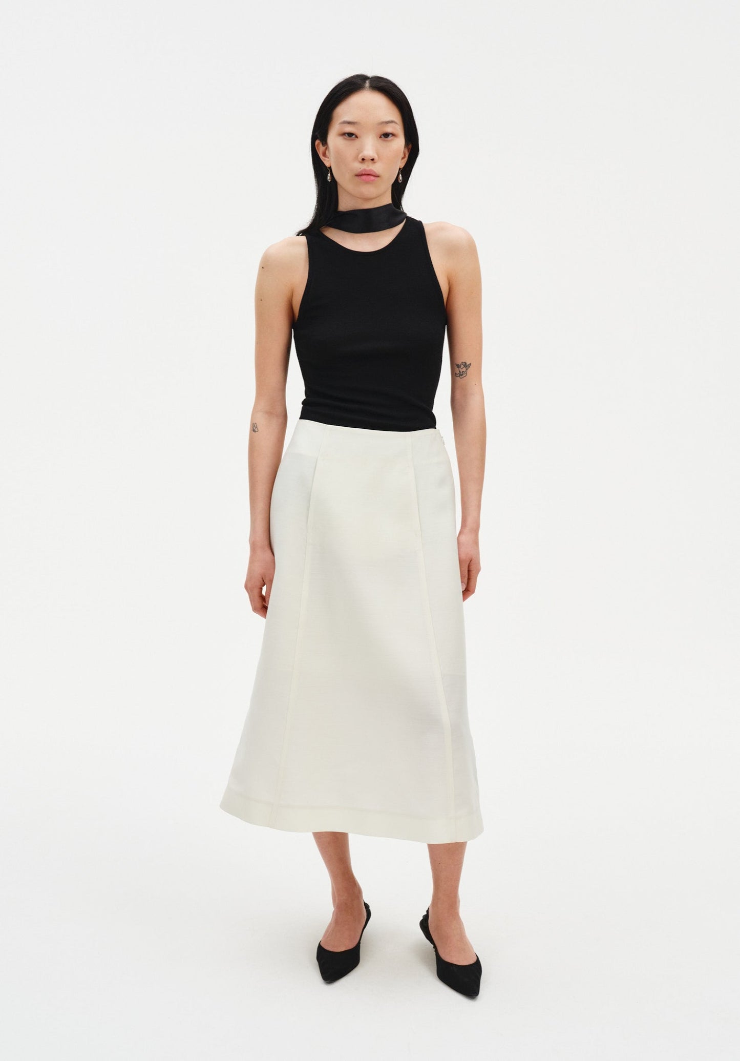 A woman stands facing forward against a plain white background, wearing a black sleeveless top with a high neckline and choker-style detail, paired with a cream-colored midi skirt and black pointed heels. Her dark shoulder-length hair is worn down, and she maintains a neutral expression with arms relaxed at her sides.
