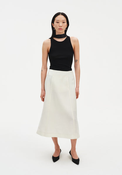A woman stands facing forward against a plain white background, wearing a black sleeveless top with a high neckline and choker-style detail, paired with a cream-colored midi skirt and black pointed heels. Her dark shoulder-length hair is worn down, and she maintains a neutral expression with arms relaxed at her sides.
